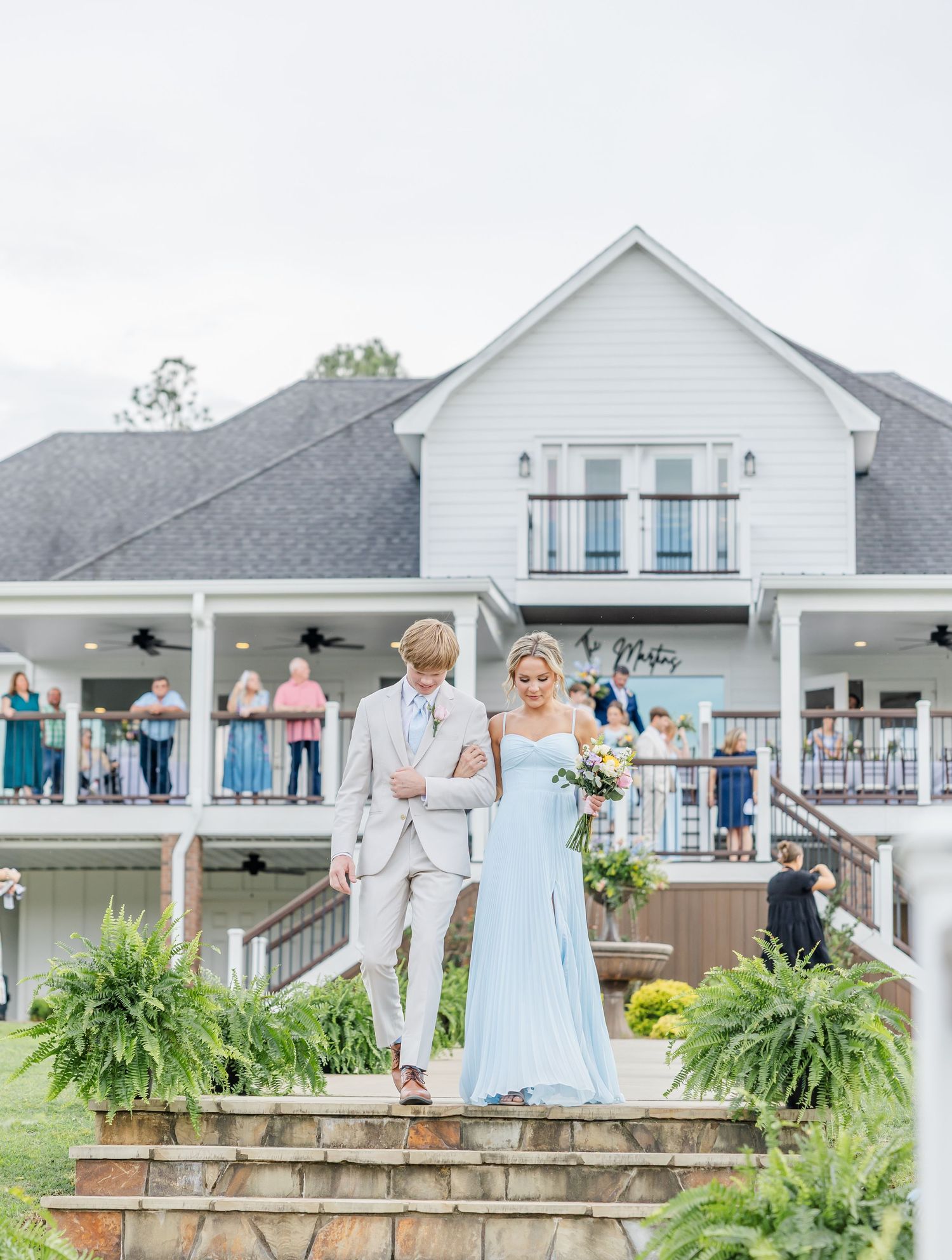 Bride in light blue gown and groom in beige suit descend stone steps at white venue.