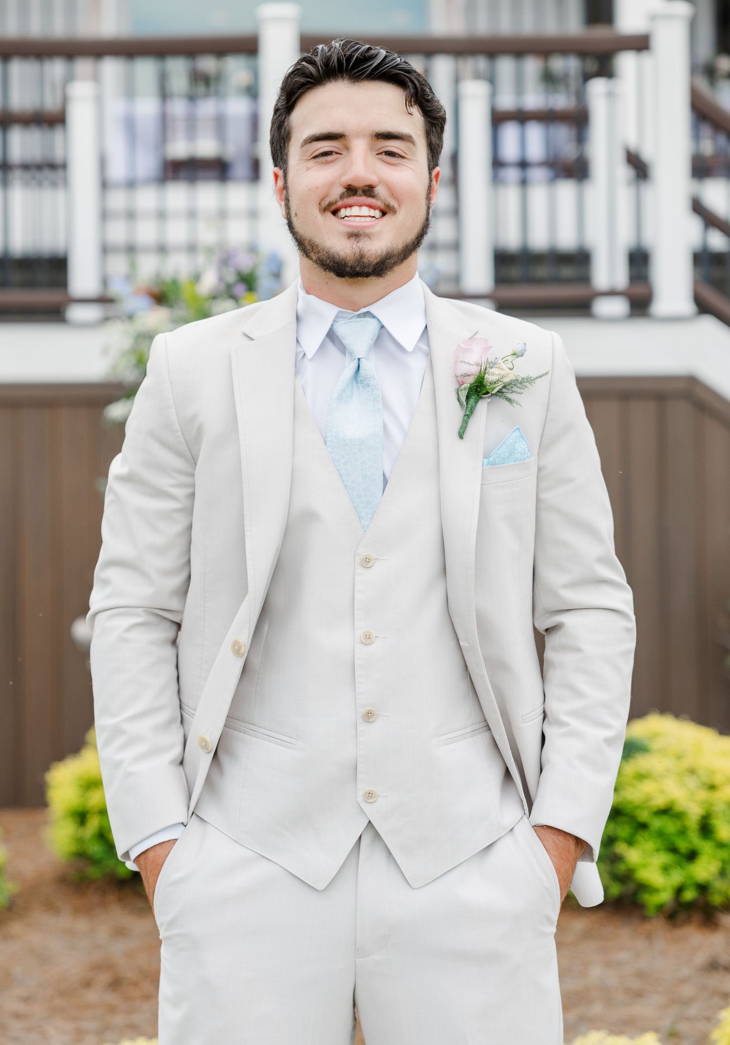Handsome groom in light beige three-piece suit with blue tie and floral boutonniere smiling outdoors.