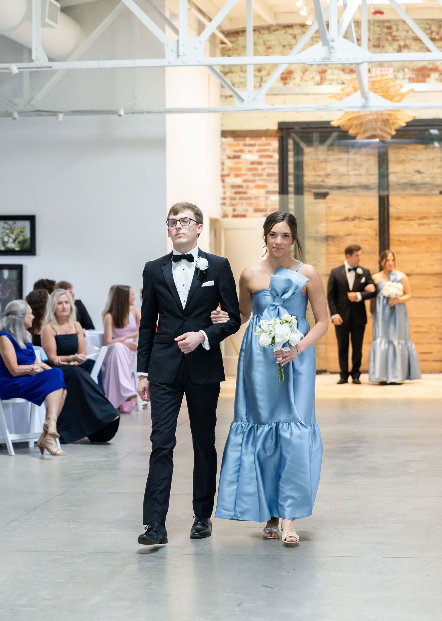 Bridesmaid in dusty blue gown walks down the aisle escorted by a groomsman in a black tuxedo.