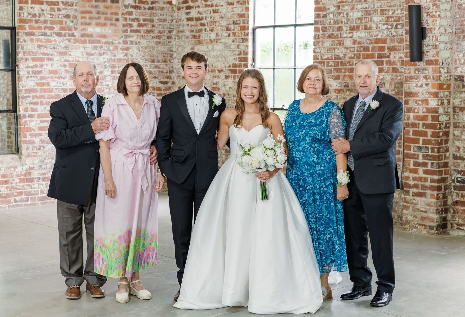Wedding party family portrait with bride, groom, and four family members posing in front of exposed brick wall.