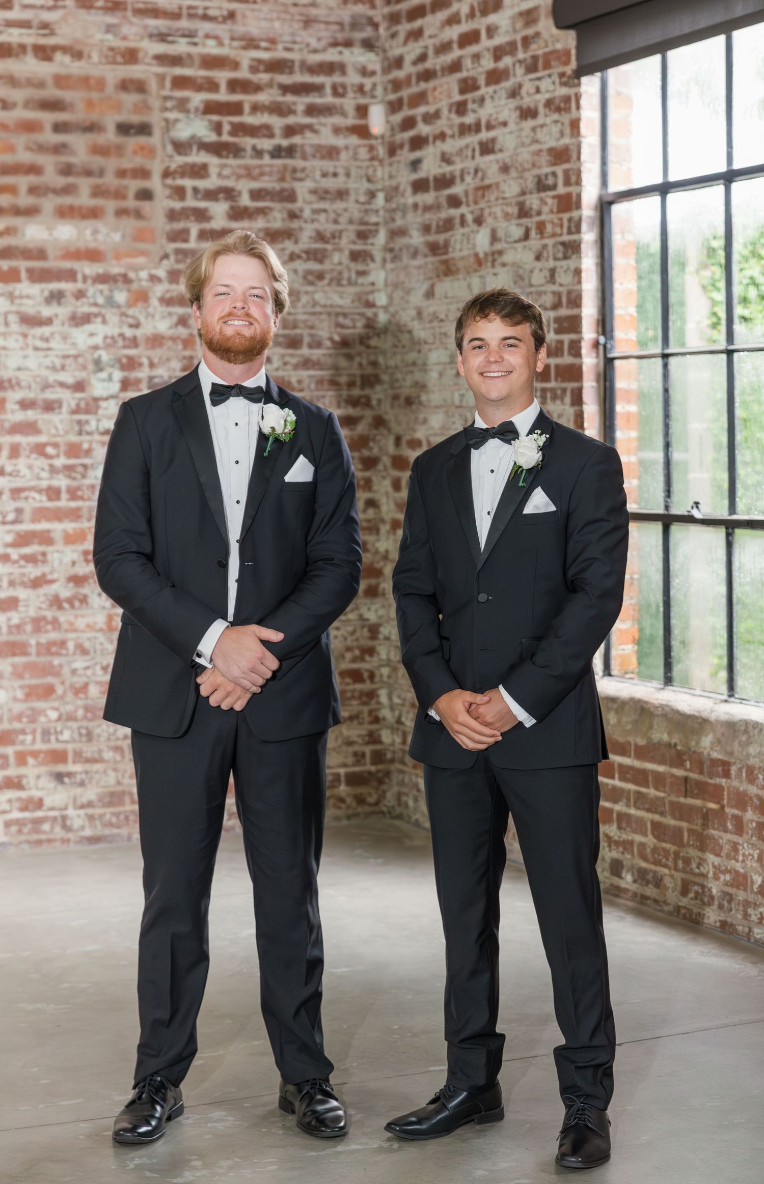 Two groomsmen in classic black tuxedos with white boutonnieres standing before exposed brick wall and large window.