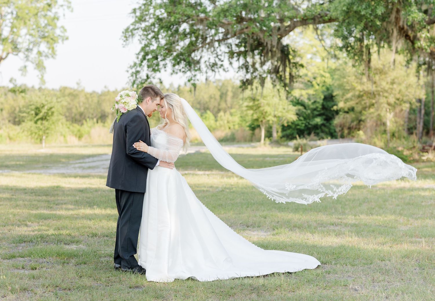 Bride and groom share romantic moment outdoors, her veil flowing in the wind, surrounded by lush greenery on their wedding day.