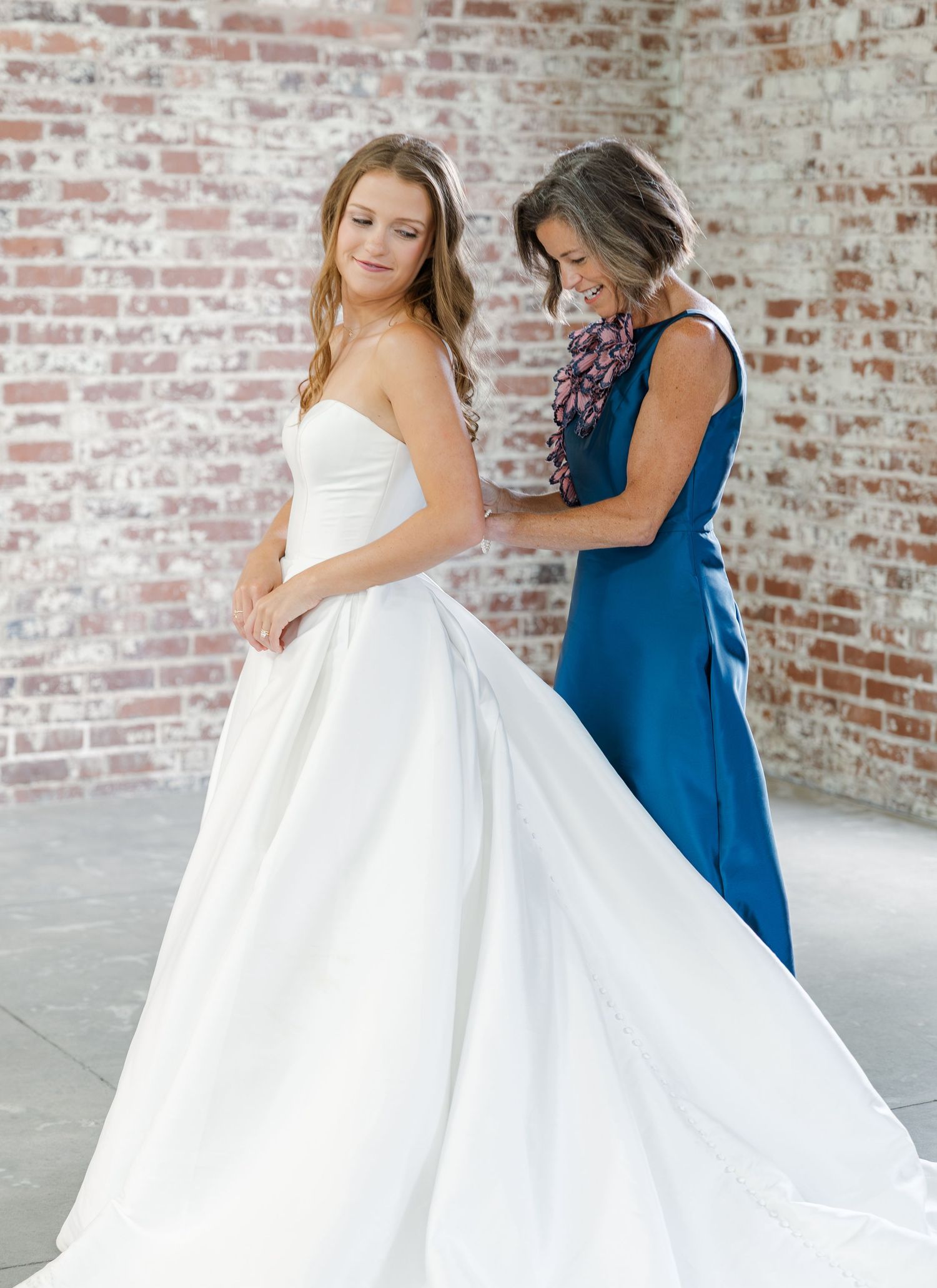 Bride in elegant strapless white ball gown being helped by woman in blue dress against exposed brick wall.