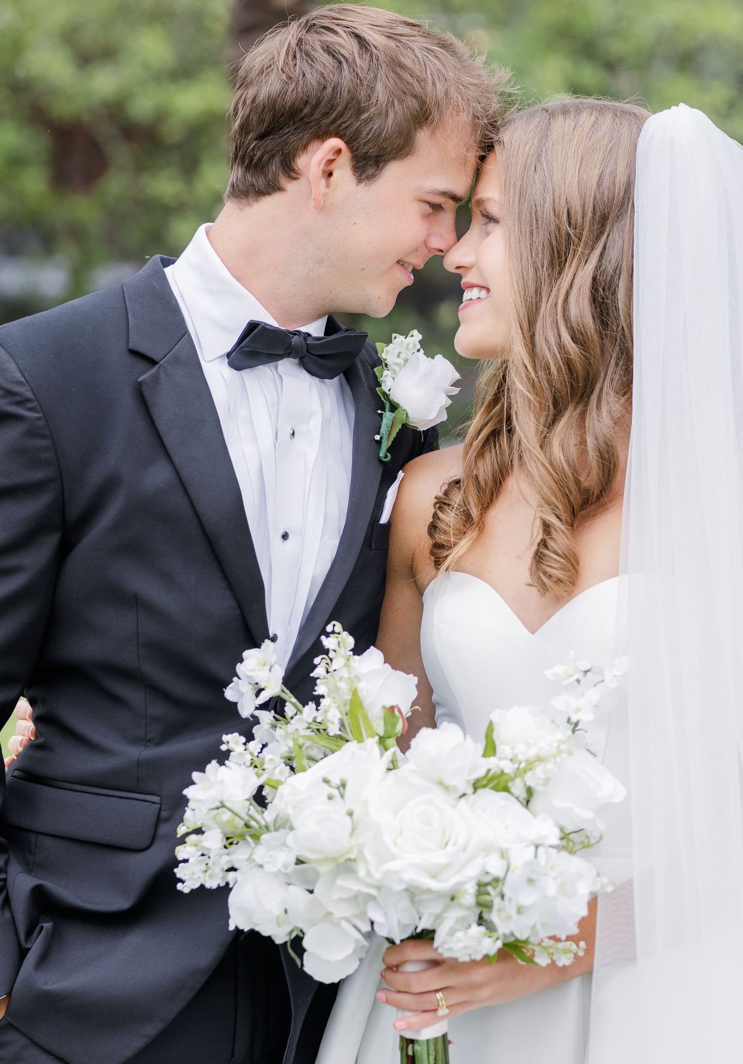 Bride and groom touching foreheads outdoors, bride holding a lush white floral bouquet, groom in black tuxedo.