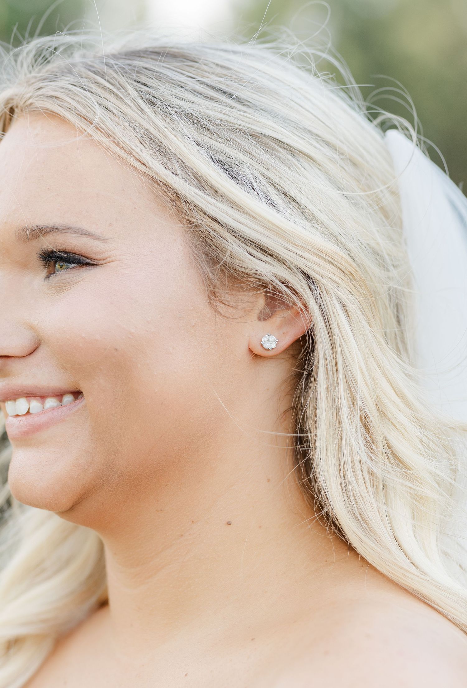 Close-up of smiling blonde bride wearing diamond stud earring and white veil on her wedding day.