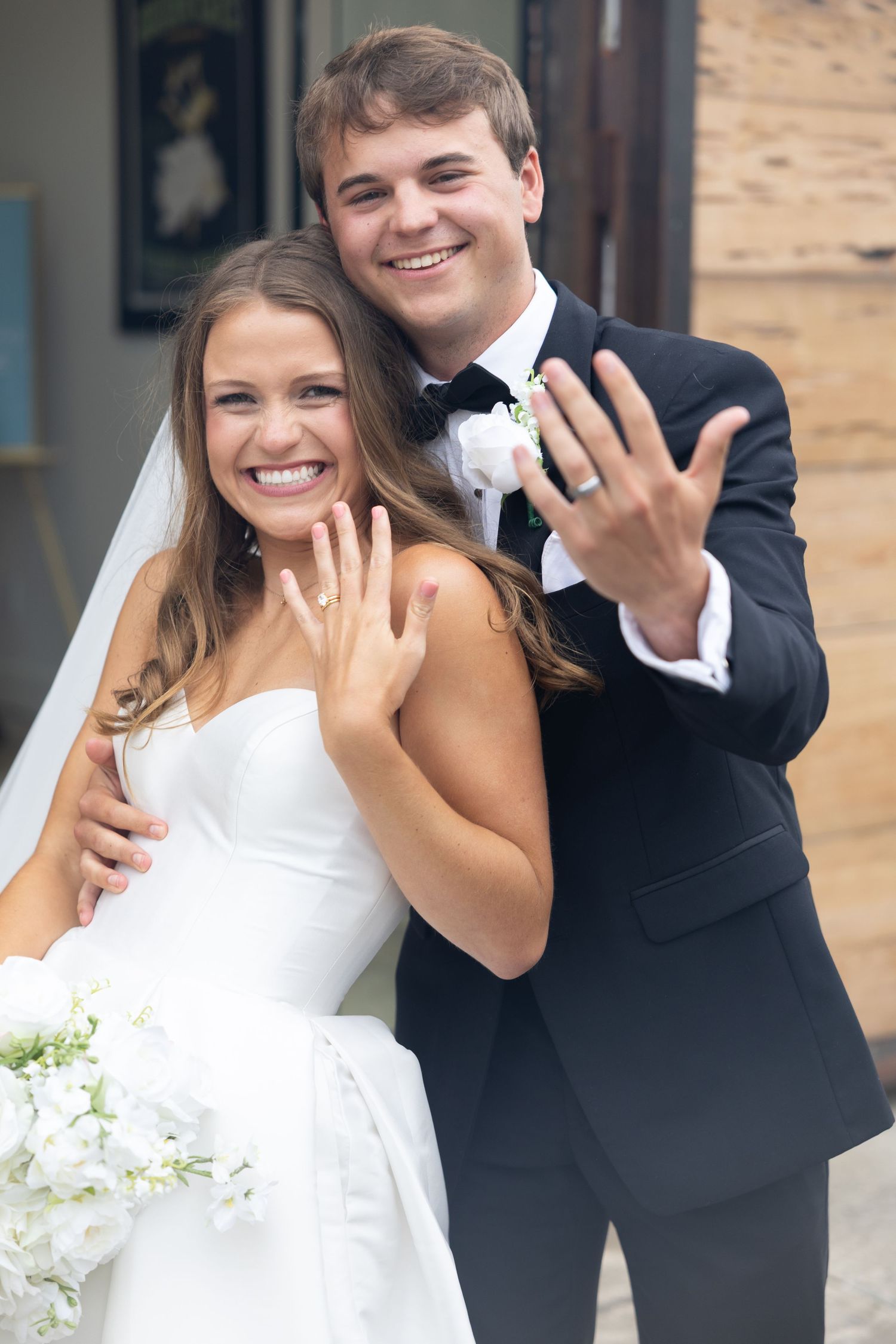 Newlyweds joyfully display wedding rings, bride in strapless gown and groom in black tuxedo.