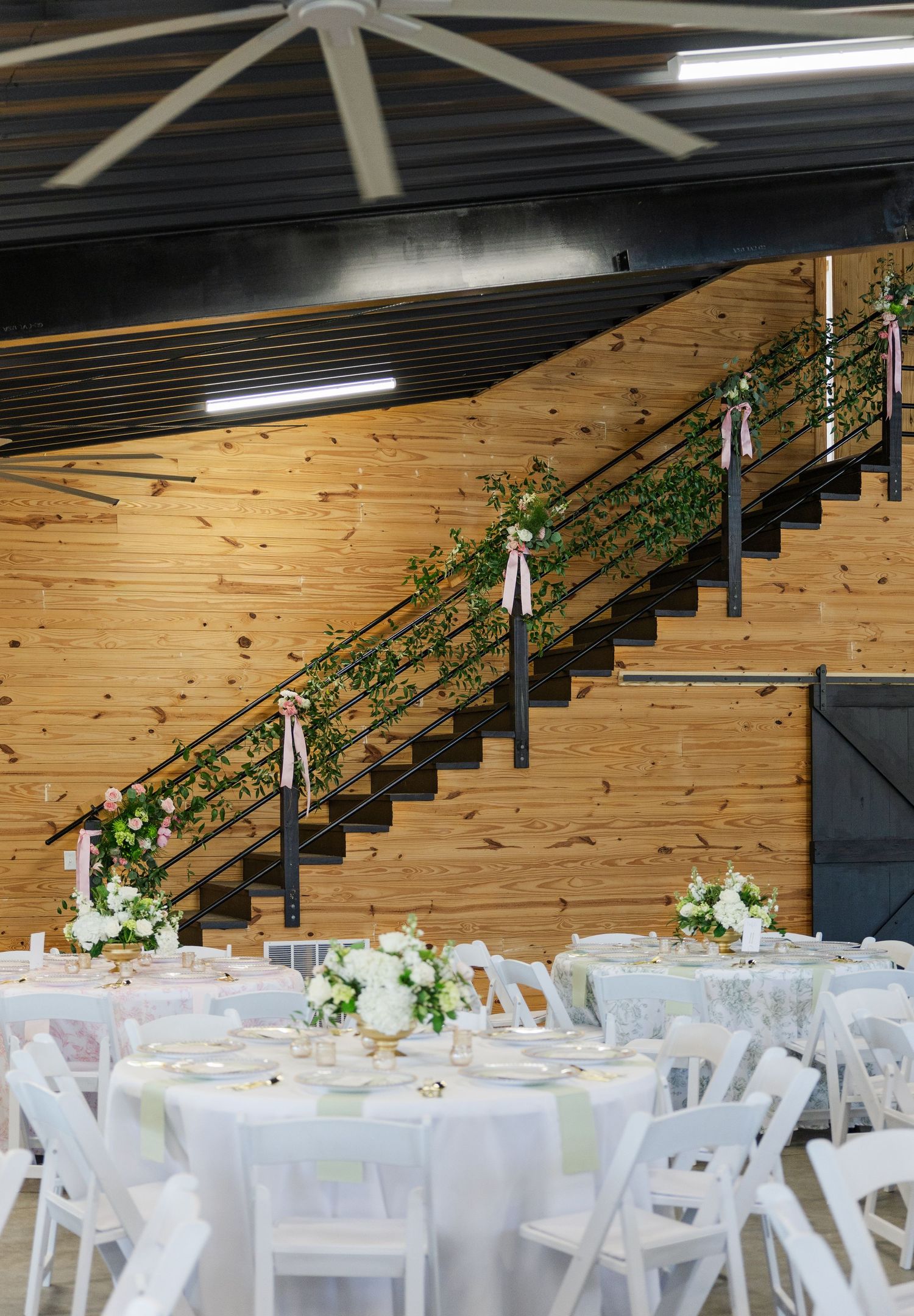 Rustic barn wedding reception with white-draped round tables, floral staircase garland with pink ribbons, and wood-paneled walls.