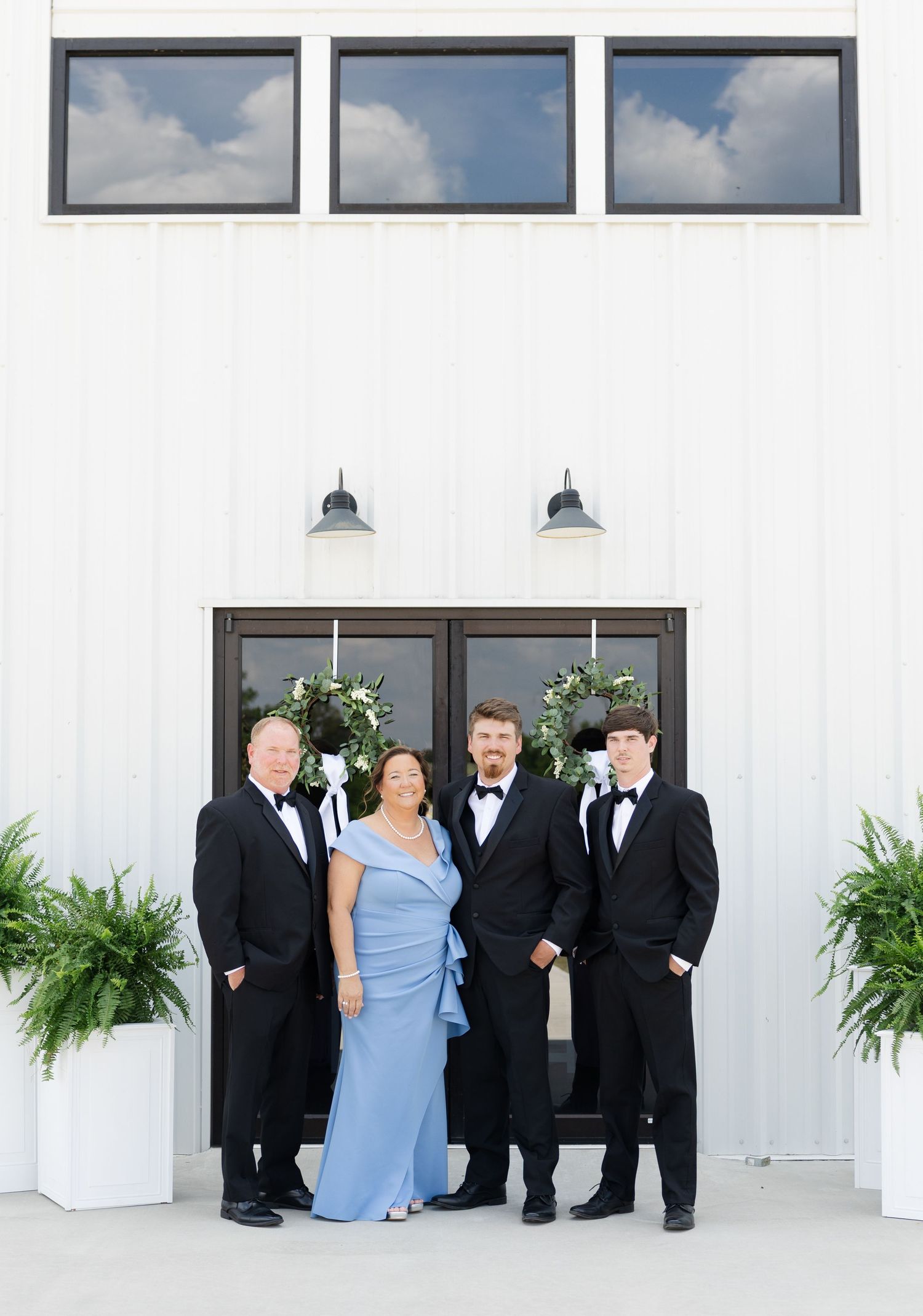 Wedding family portrait with four members in formal attire outside a white barn venue with floral wreaths.