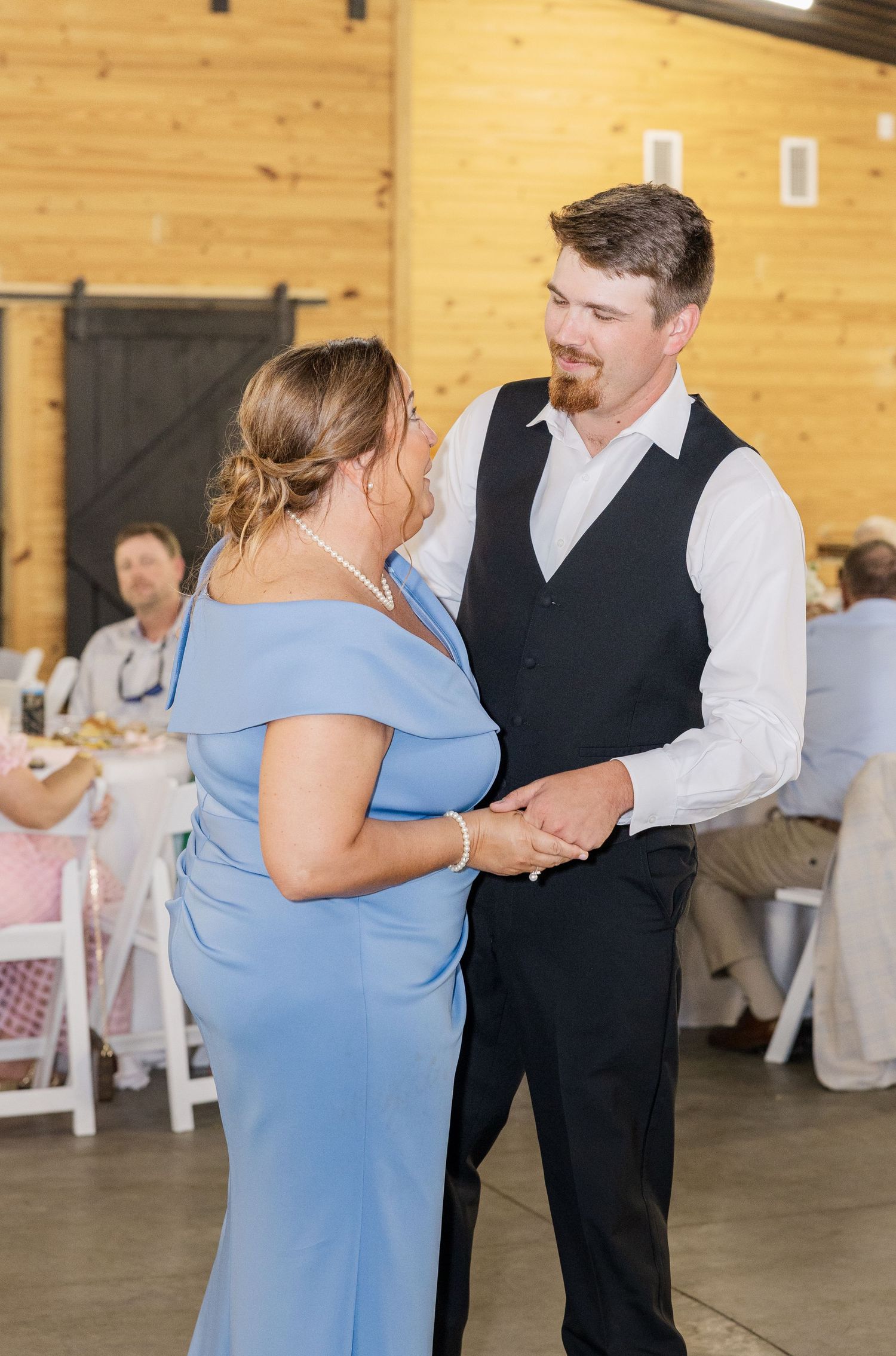 Groom in black vest dances with woman in blue off-shoulder gown at rustic barn wedding reception.