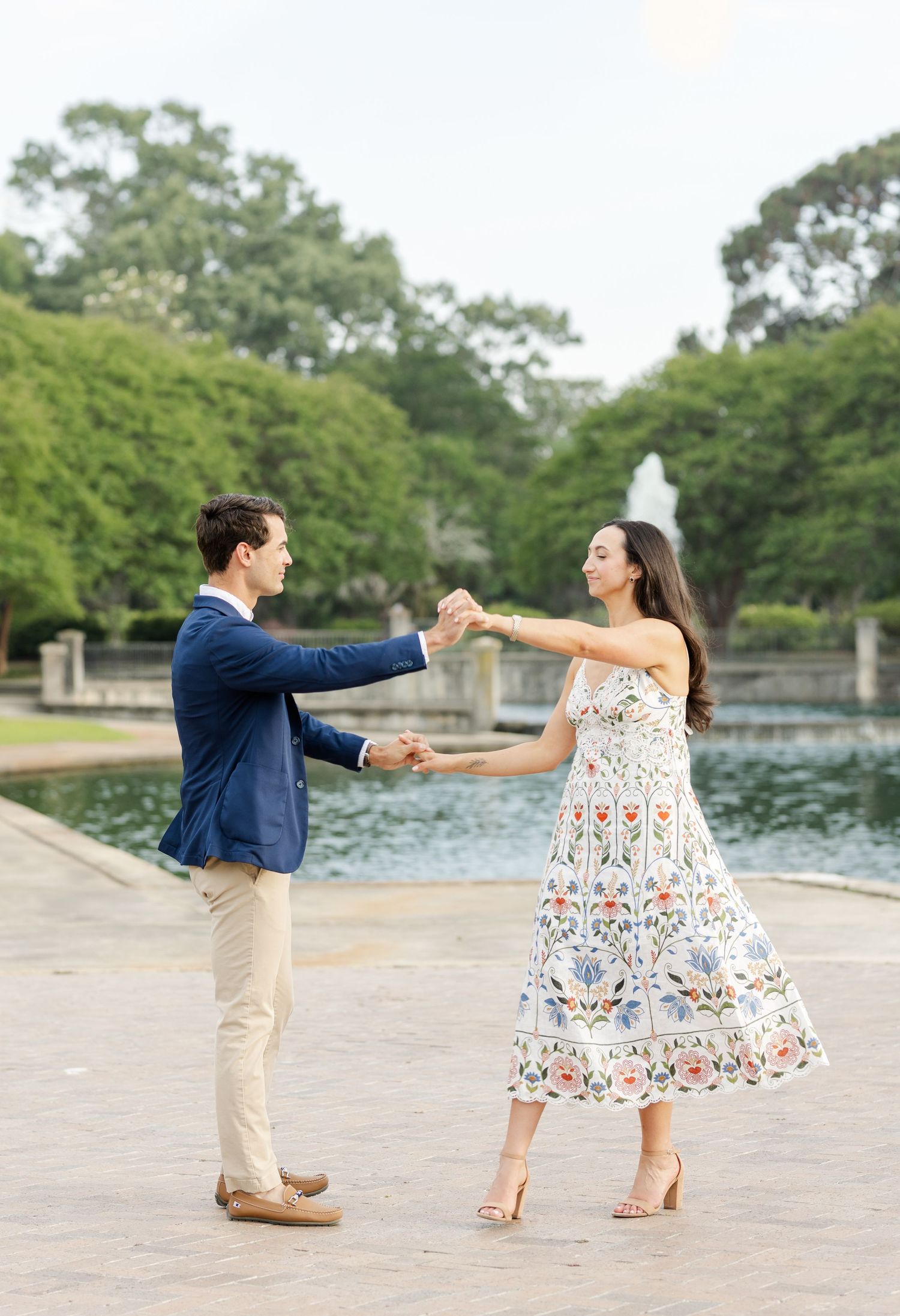 A couple dancing joyfully by a fountain in a park, woman wearing a floral midi dress.