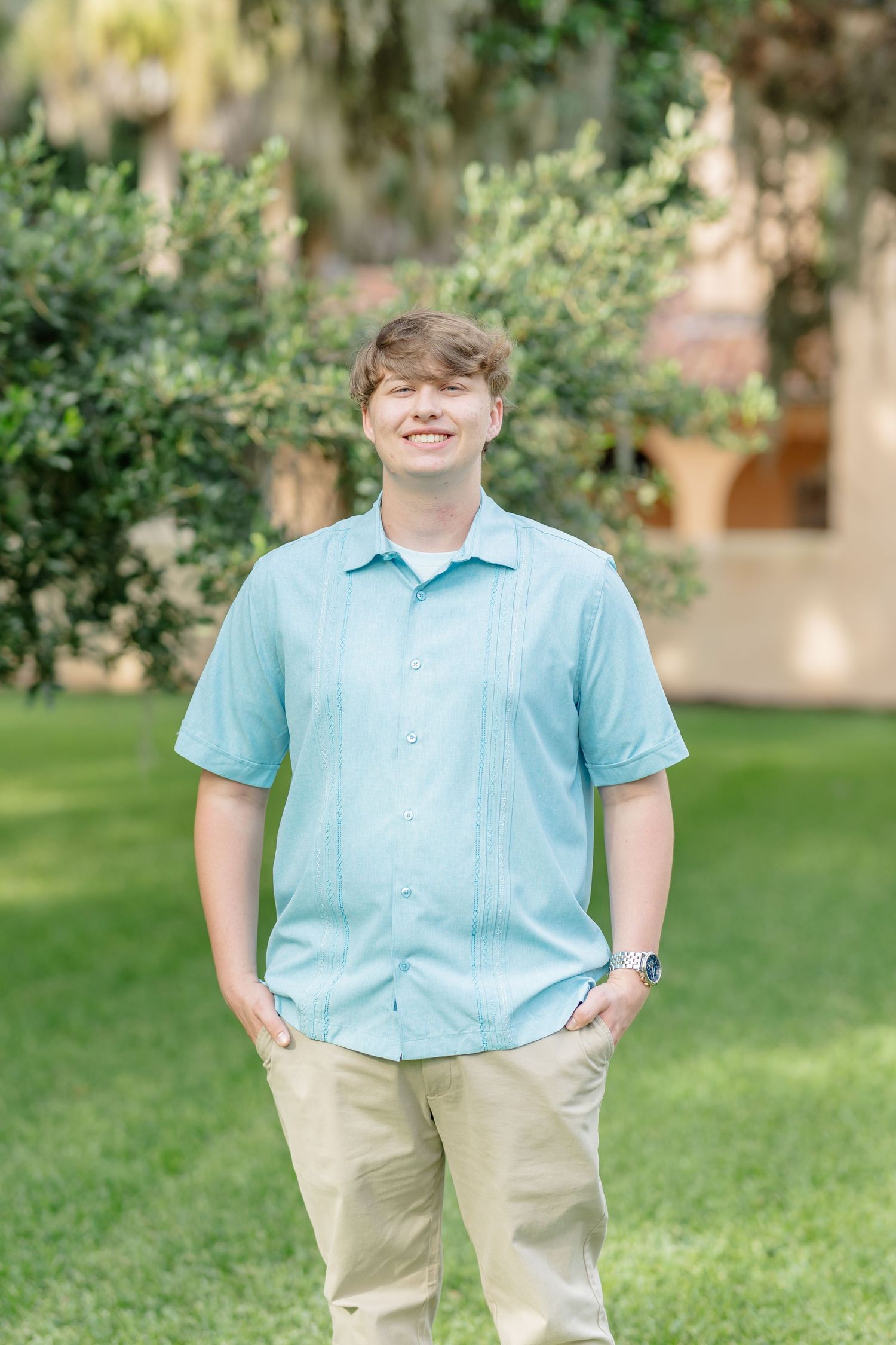 Someone wearing a light blue shirt and khaki pants poses casually in an outdoor garden setting.