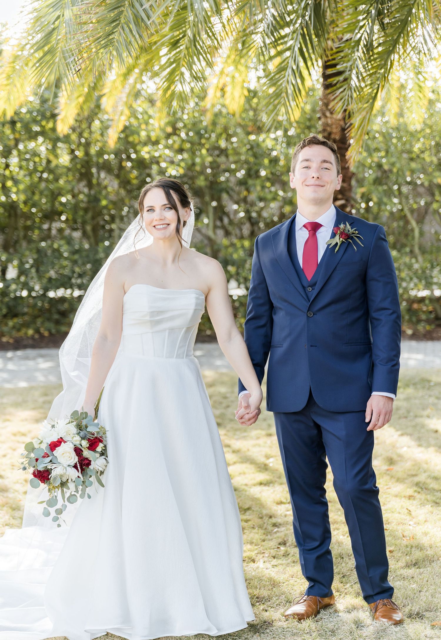 A wedding couple poses under palm trees in a sunny garden setting.