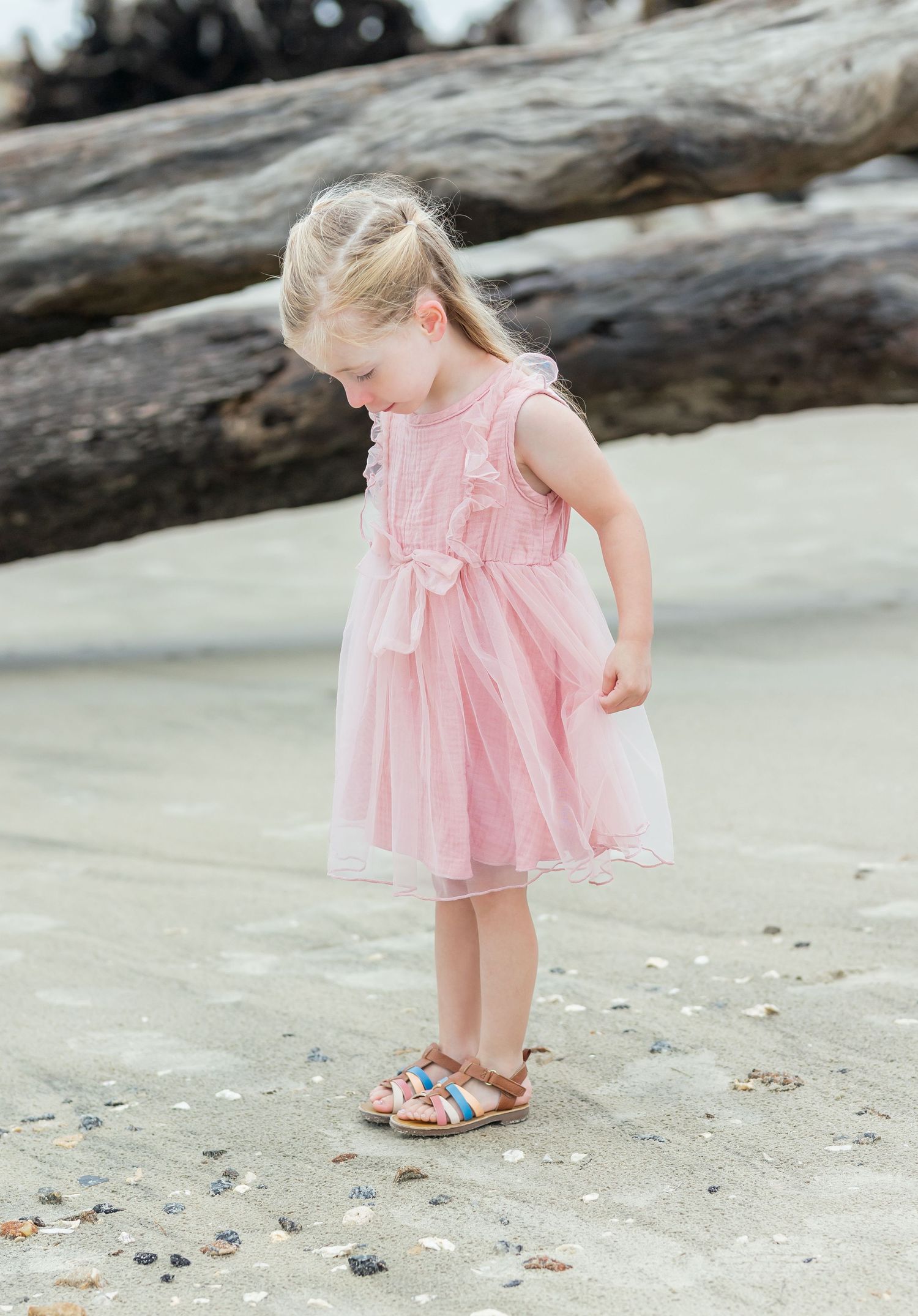 A young child in a pink ruffled dress explores seashells and sand on a beach near driftwood.
