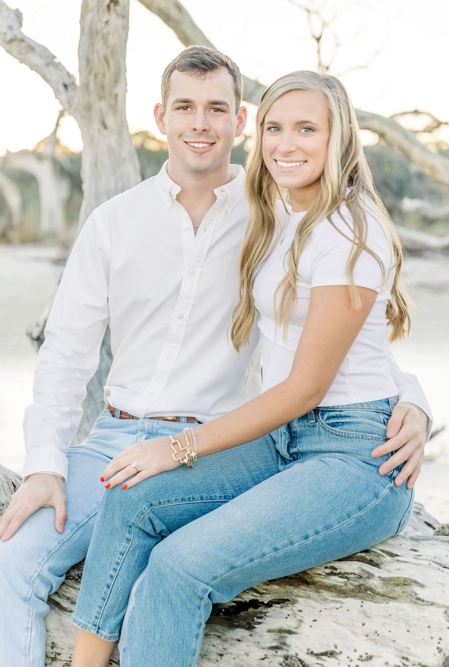 A couple in white and denim sits casually together on a beach during a bright sunny day.