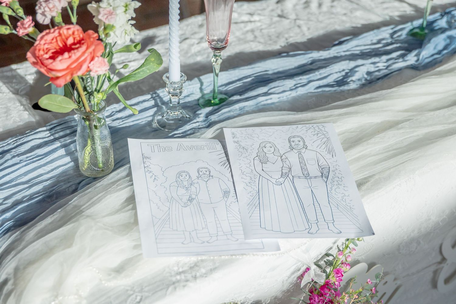 Wedding sketch illustrations displayed on a white tablecloth with pink flowers in a vase and champagne glasses.