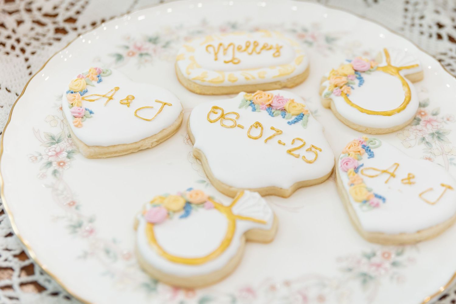 Elegant white sugar cookies with gold trim and wedding details displayed on a floral-patterned plate.