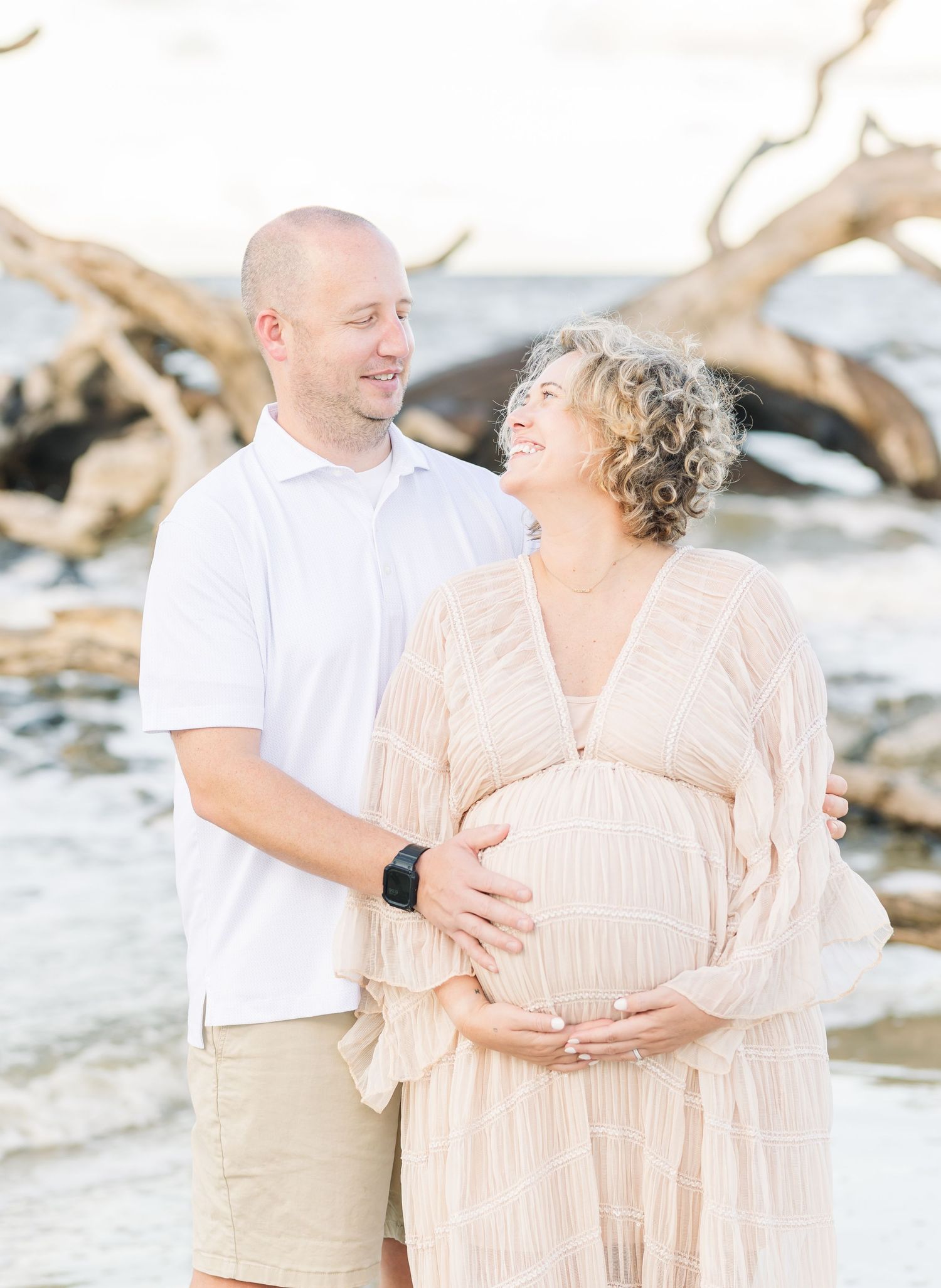 Romantic maternity photoshoot on beach with driftwood backdrop and soft lighting at sunset.