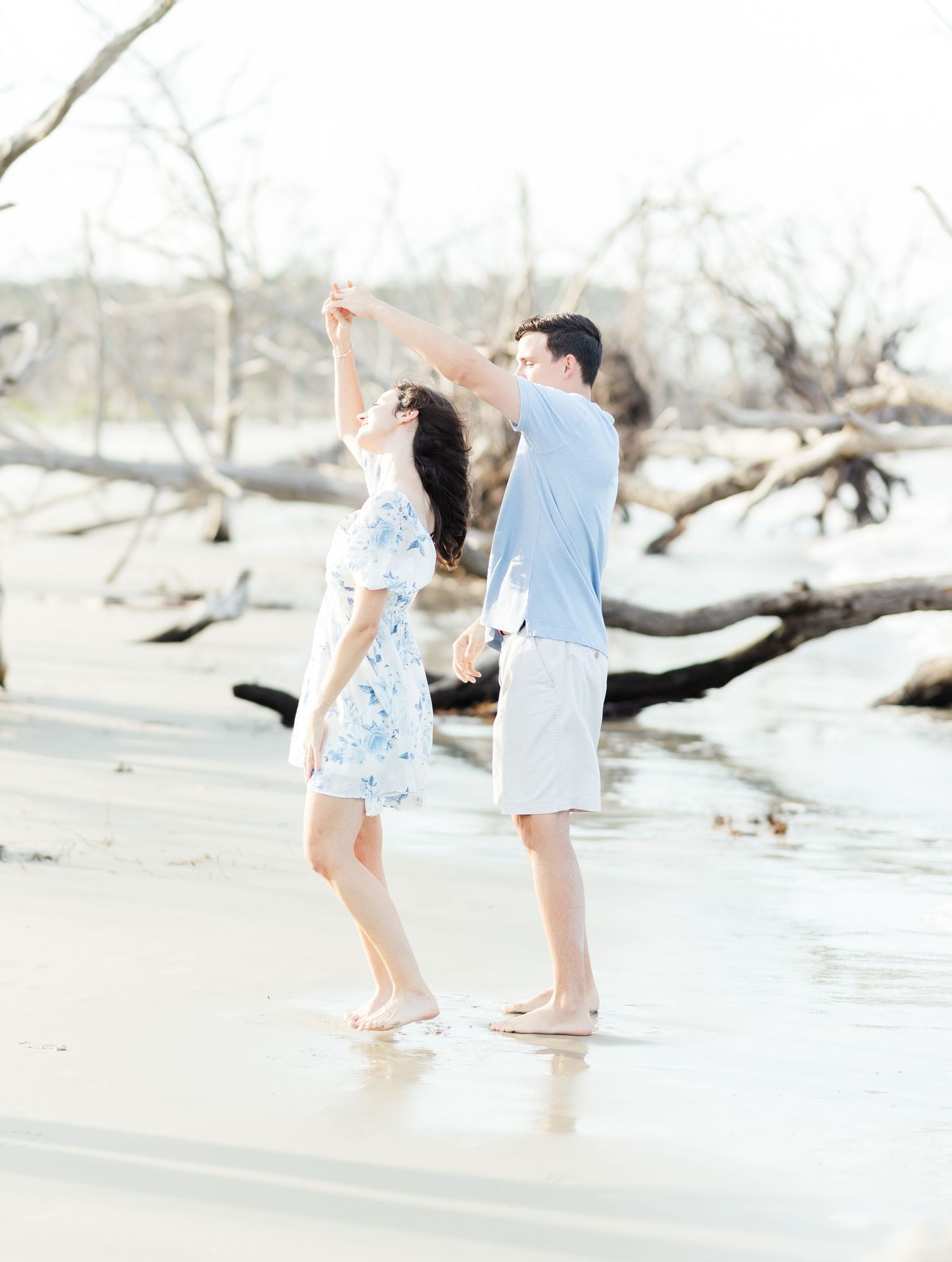 A playful couple dances together on a serene beach with driftwood scattered nearby.