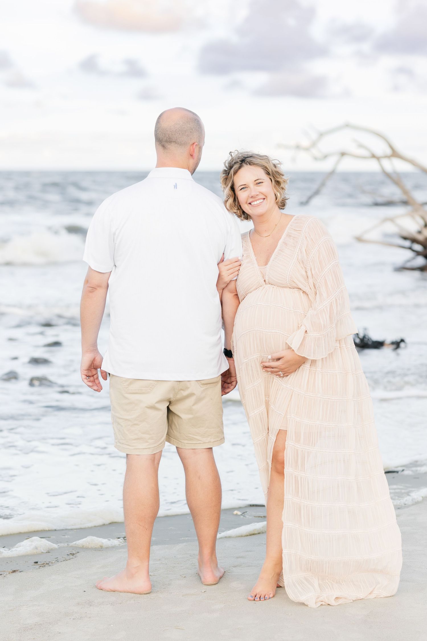 A romantic beach photoshoot featuring a couple standing barefoot in the sand by the ocean waves.