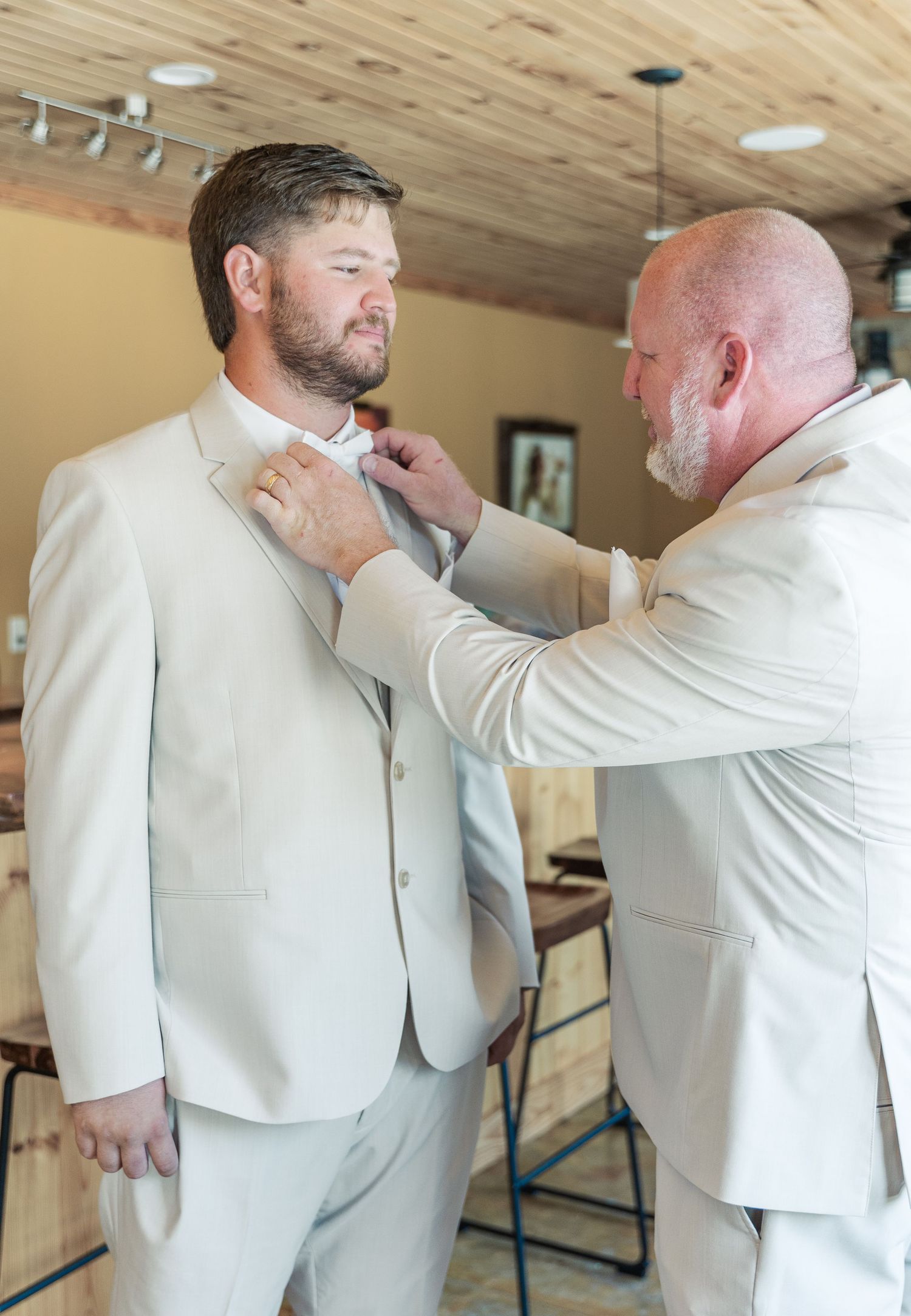 A groom gets help adjusting his tie before the wedding ceremony in a rustic wood-paneled room.