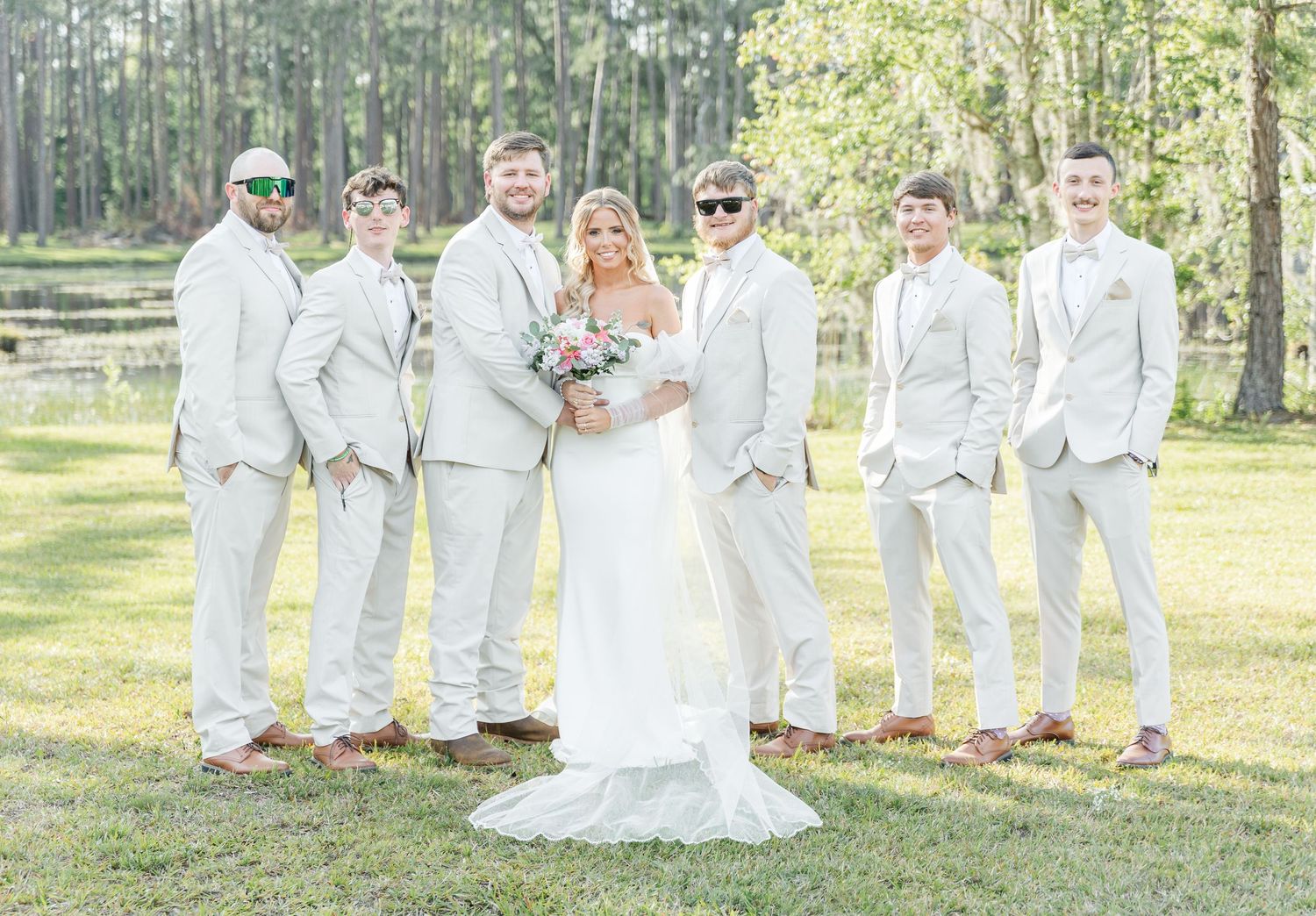 Wedding party in all-white suits poses with couple on sunny lawn.