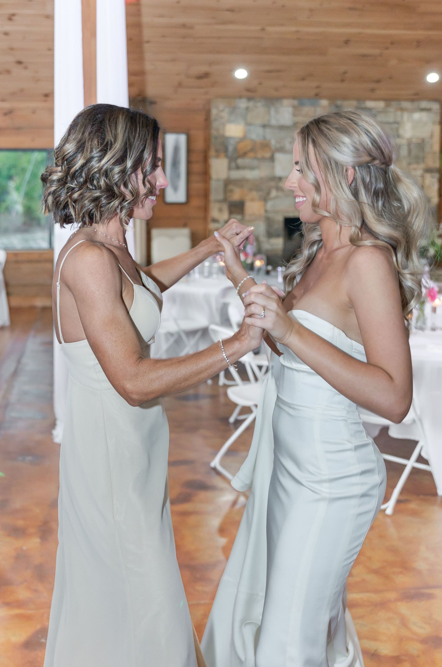Two people in light-colored dresses dancing together at a rustic wedding reception venue.