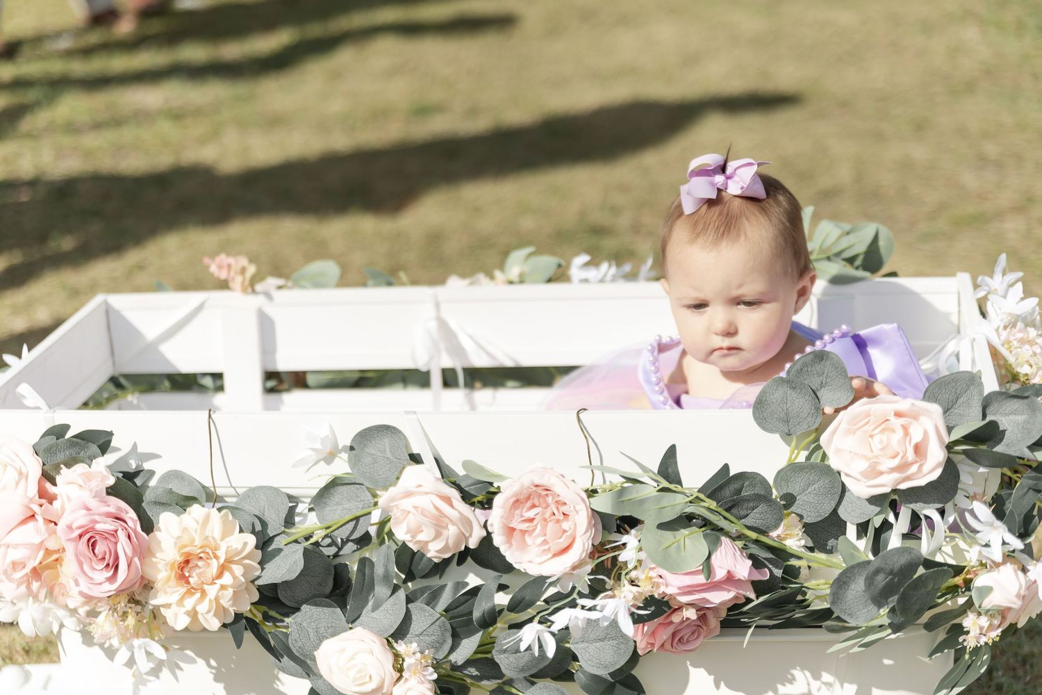 Baby in a white wagon decorated with pink roses and greenery at outdoor event.