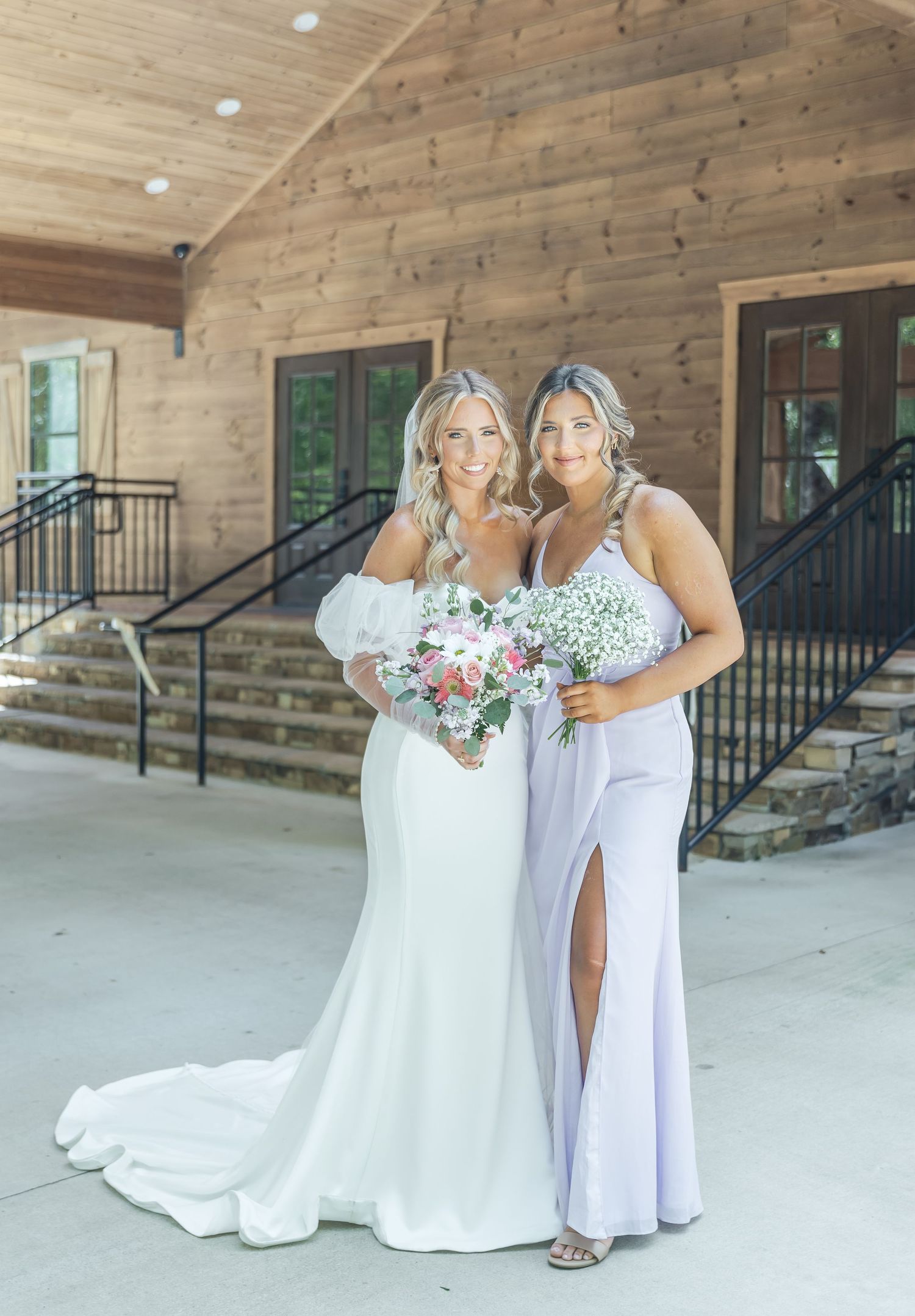 Wedding party members pose in white and lavender dresses with floral bouquets on rustic venue steps.