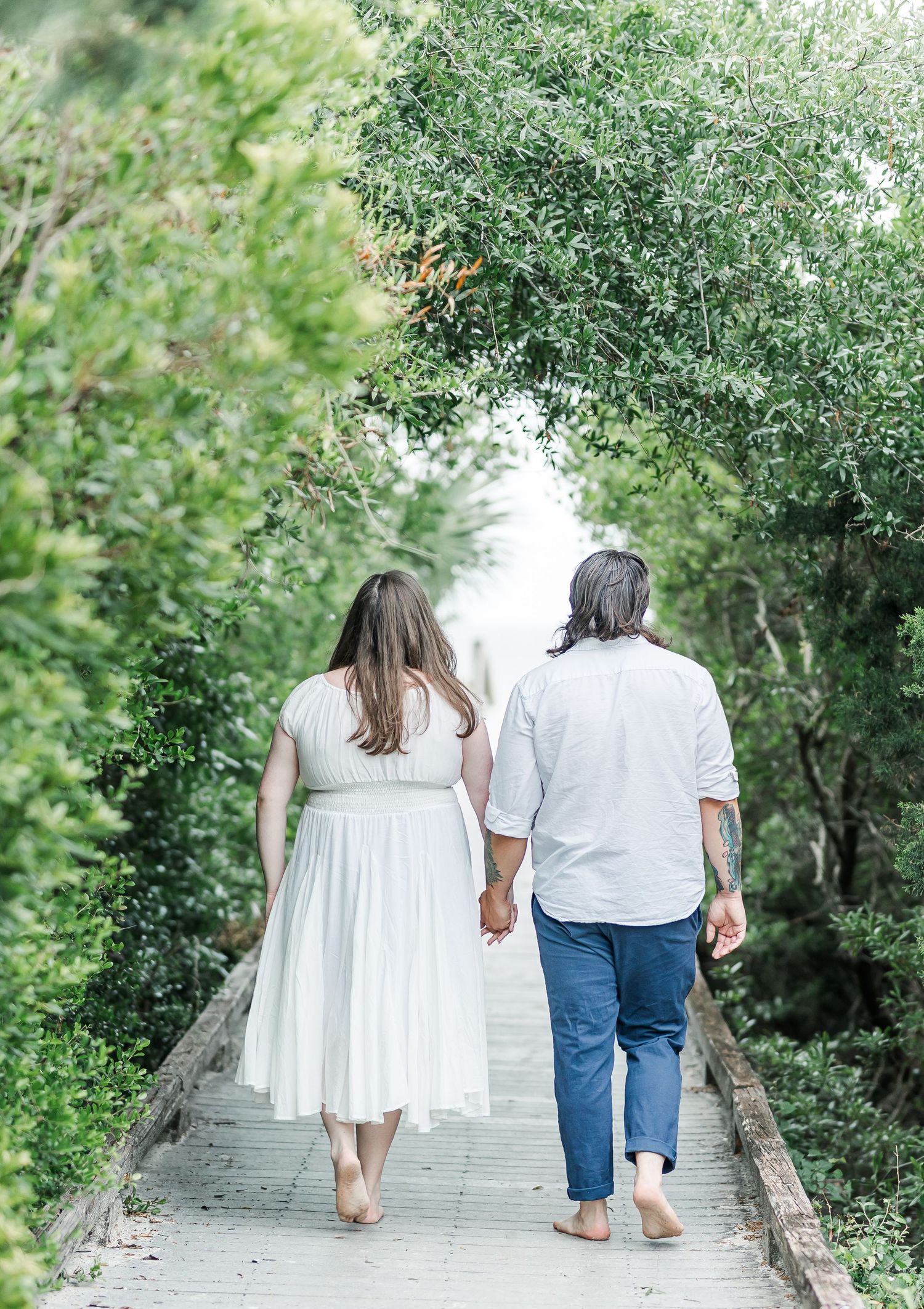 Couple walks barefoot hand in hand down a scenic wooden pathway surrounded by trees.