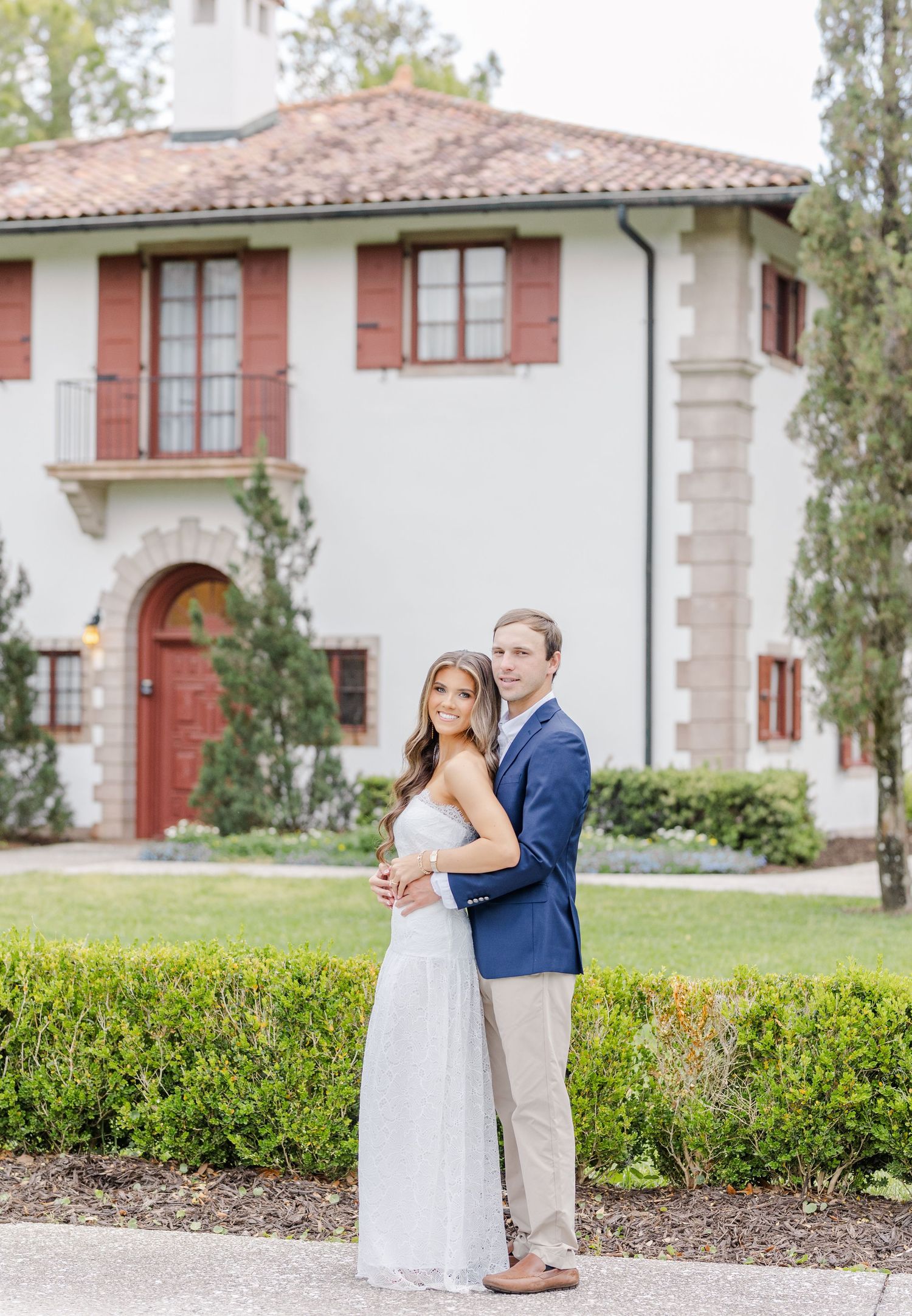 Engaged couple embraces in front of a white Spanish-style villa surrounded by lush greenery.