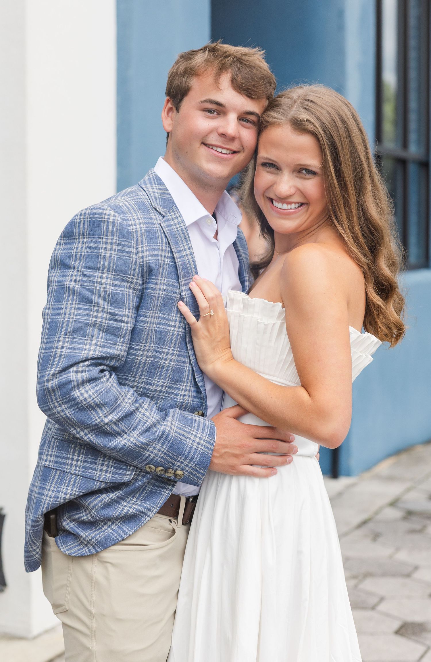 A smiling young couple poses outdoors, the man in a blue plaid blazer and the woman in a white strapless dress hugging him.