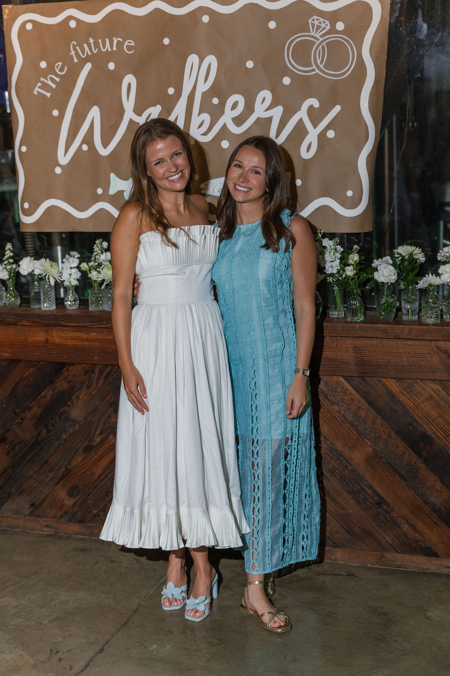 Two smiling women pose together at engagement party, one in white dress, one in teal lace dress.