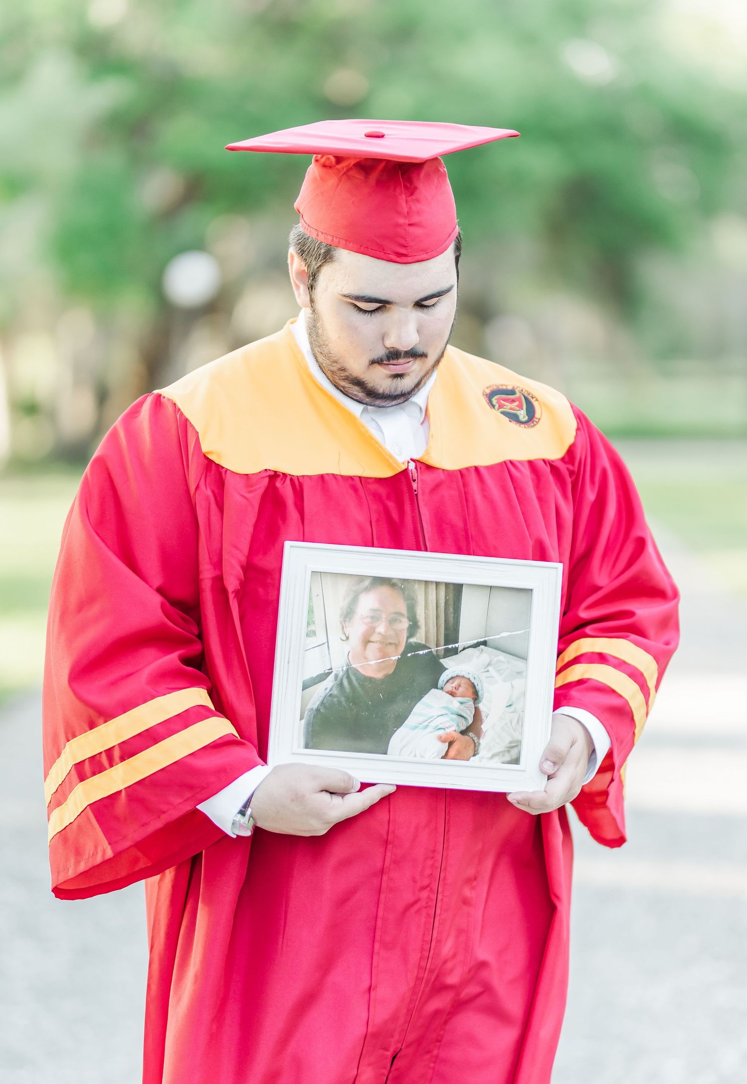 Graduate in red cap and gown holds a framed photograph while standing outdoors during commencement.