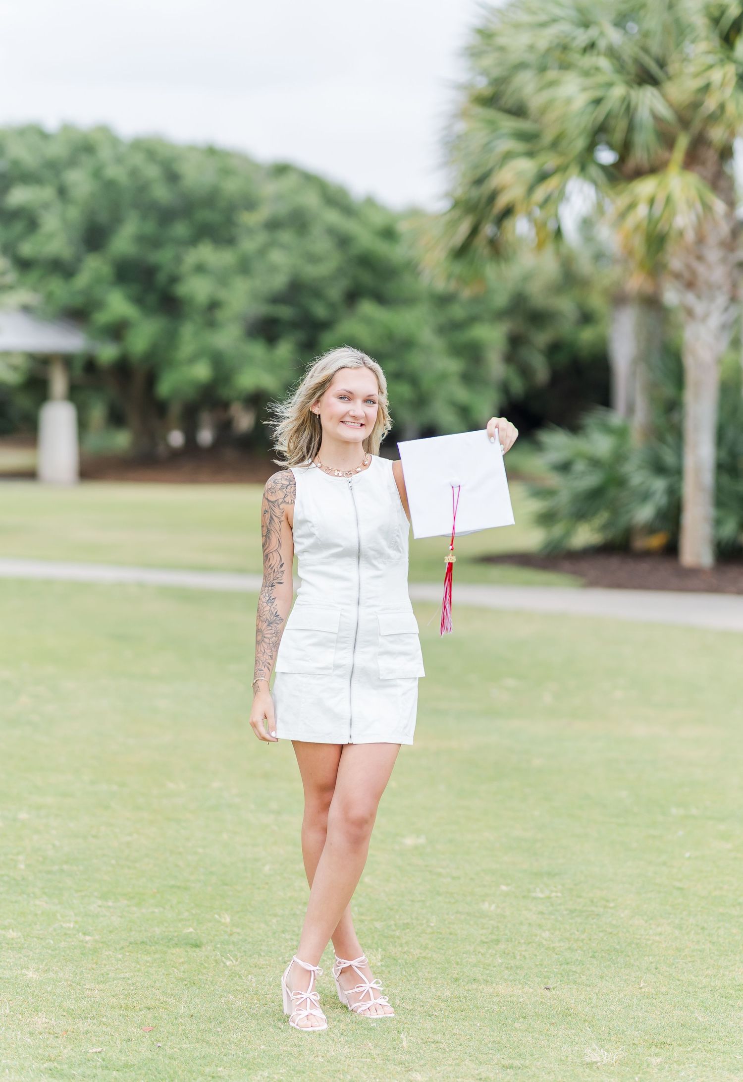 Graduate in white dress holding diploma walks across a sunny lawn with palm trees.