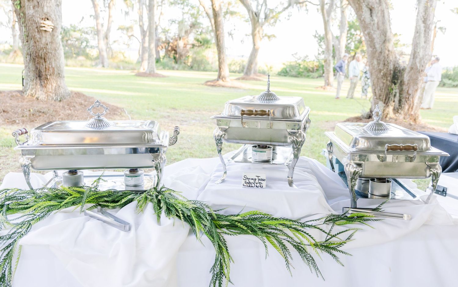 Elegant outdoor event setup with white tables and chairs arranged under trees for a garden party.