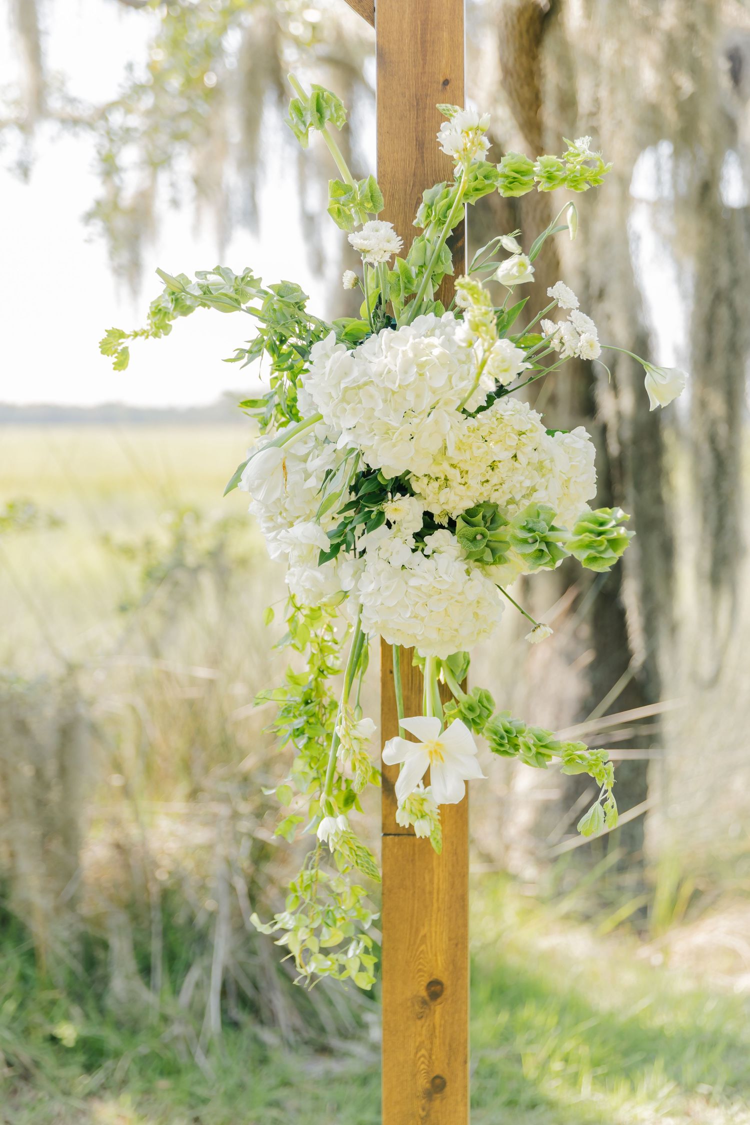 Elegant white floral arrangement with cascading greenery adorns a wooden post against a dreamy outdoor backdrop.