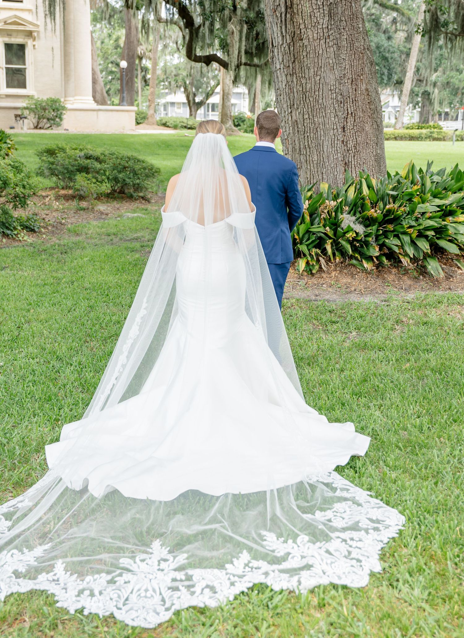 A long flowing cathedral veil with lace details trails behind a couple standing together in a lush garden setting.