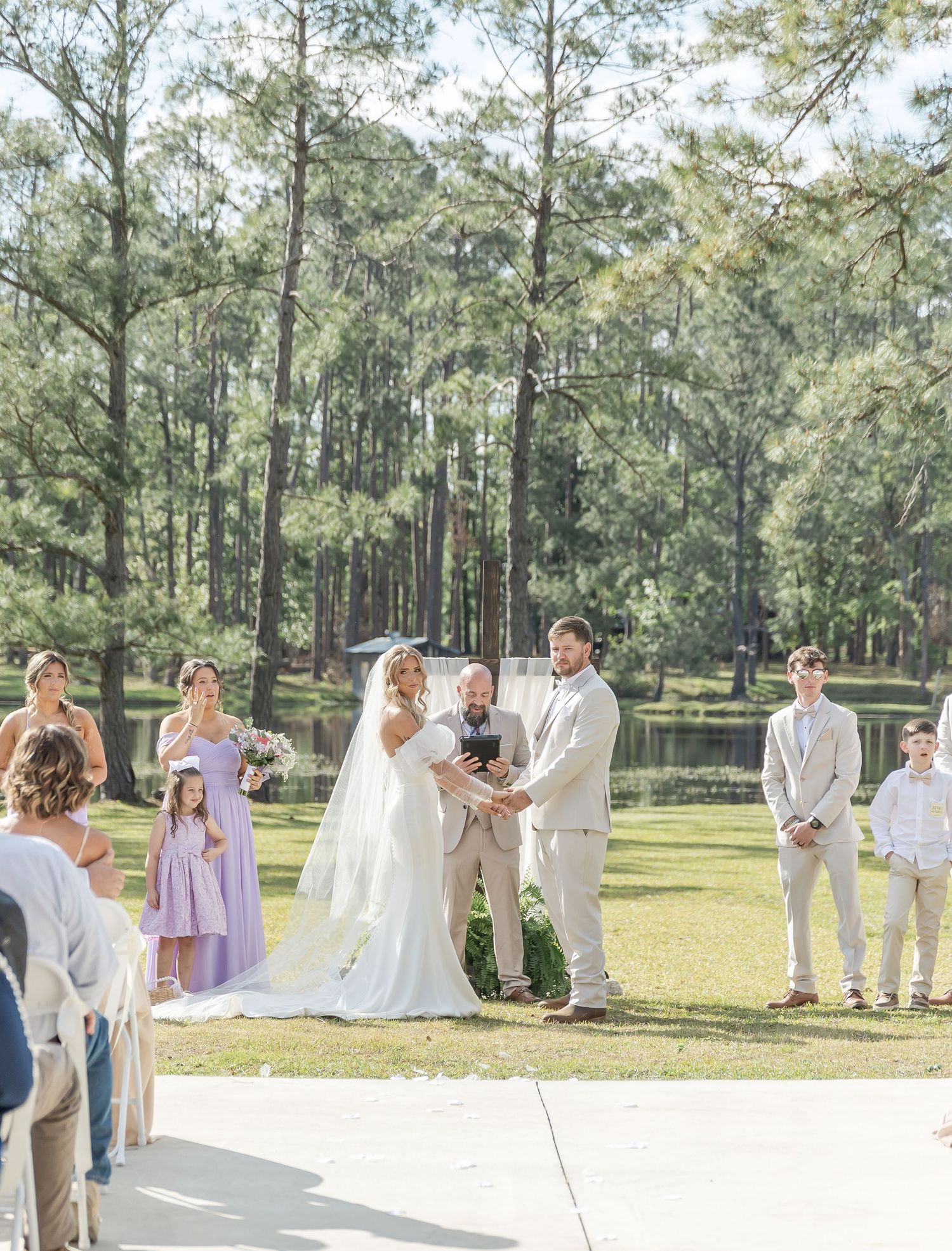 Outdoor wedding ceremony takes place with pine forest backdrop and guests seated on white chairs.