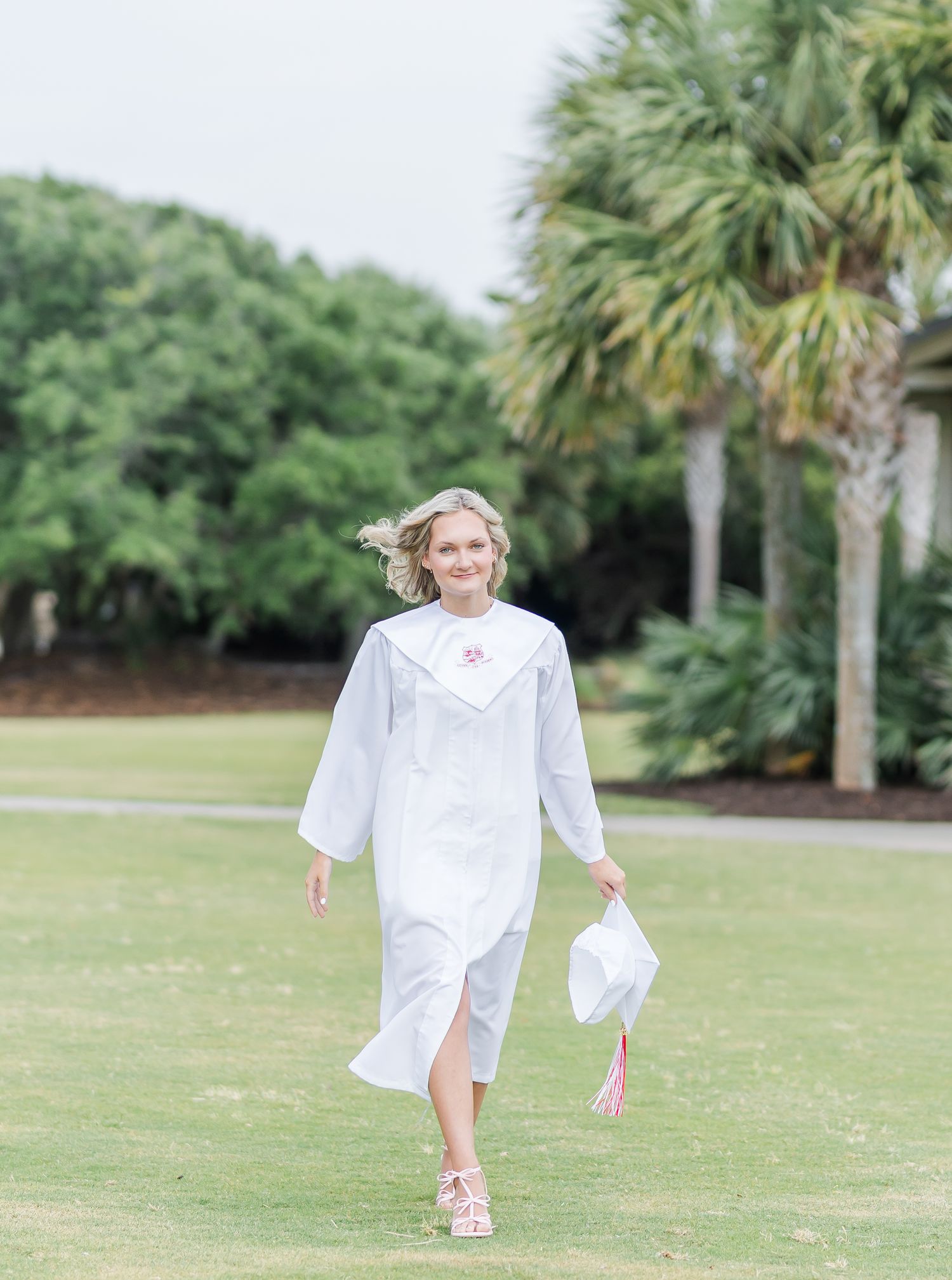 A person walks across a grassy field wearing a white graduation gown with palm trees in the background.
