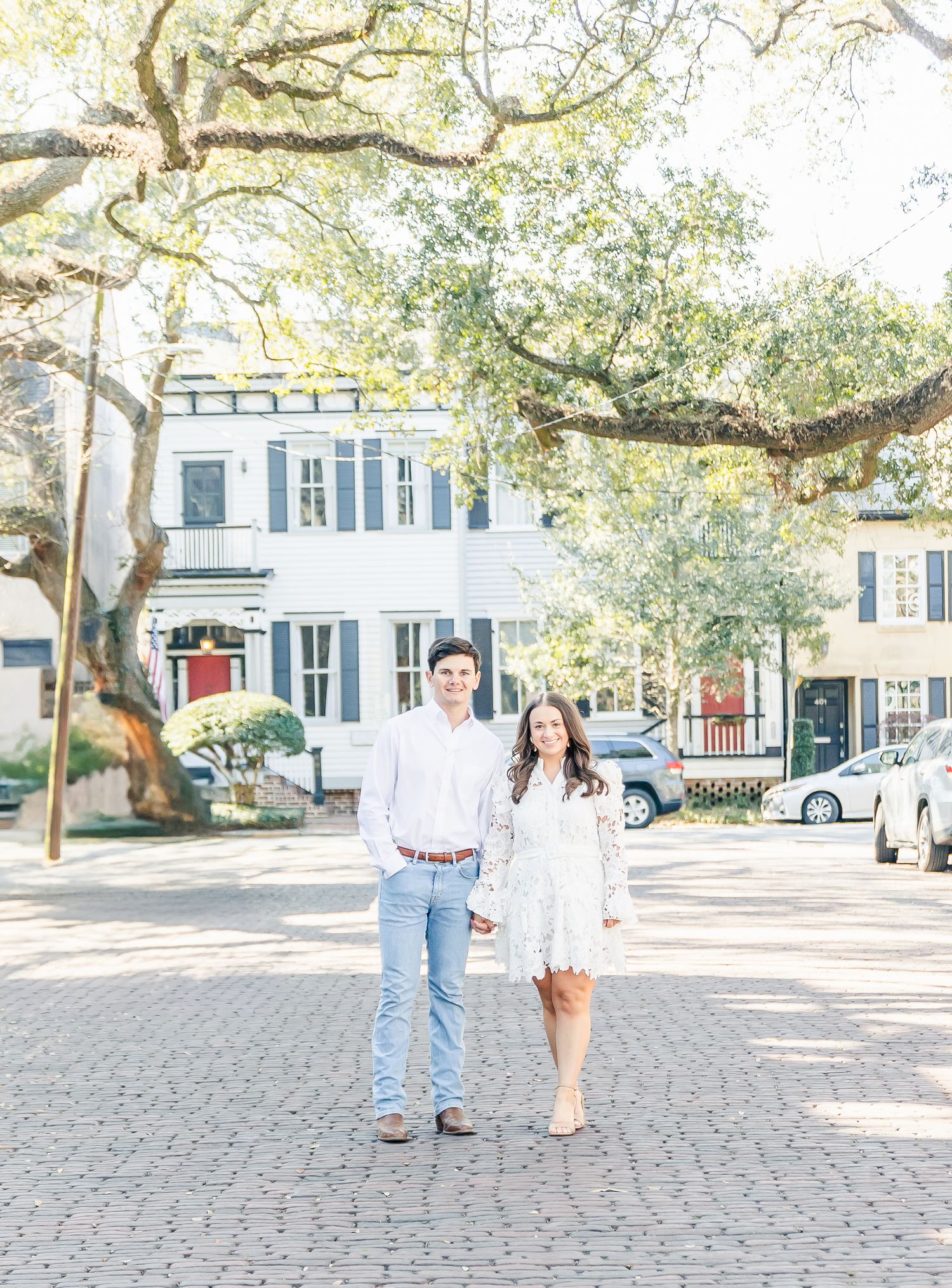 A couple stands on a cobblestone street lined with oak trees and historic homes in Charleston.