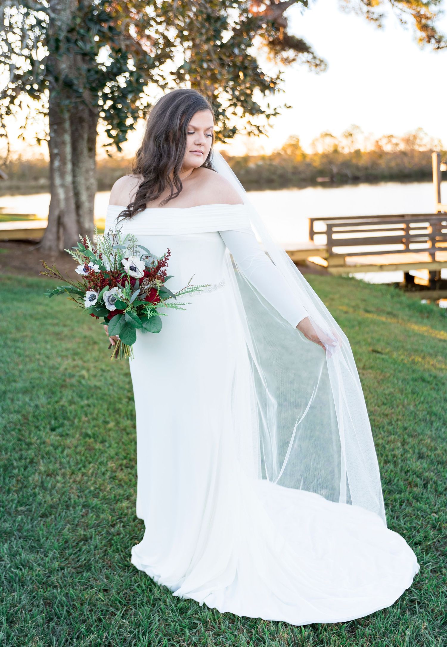 An off-shoulder white wedding dress with flowing sleeves and train photographed at sunset by a lake.
