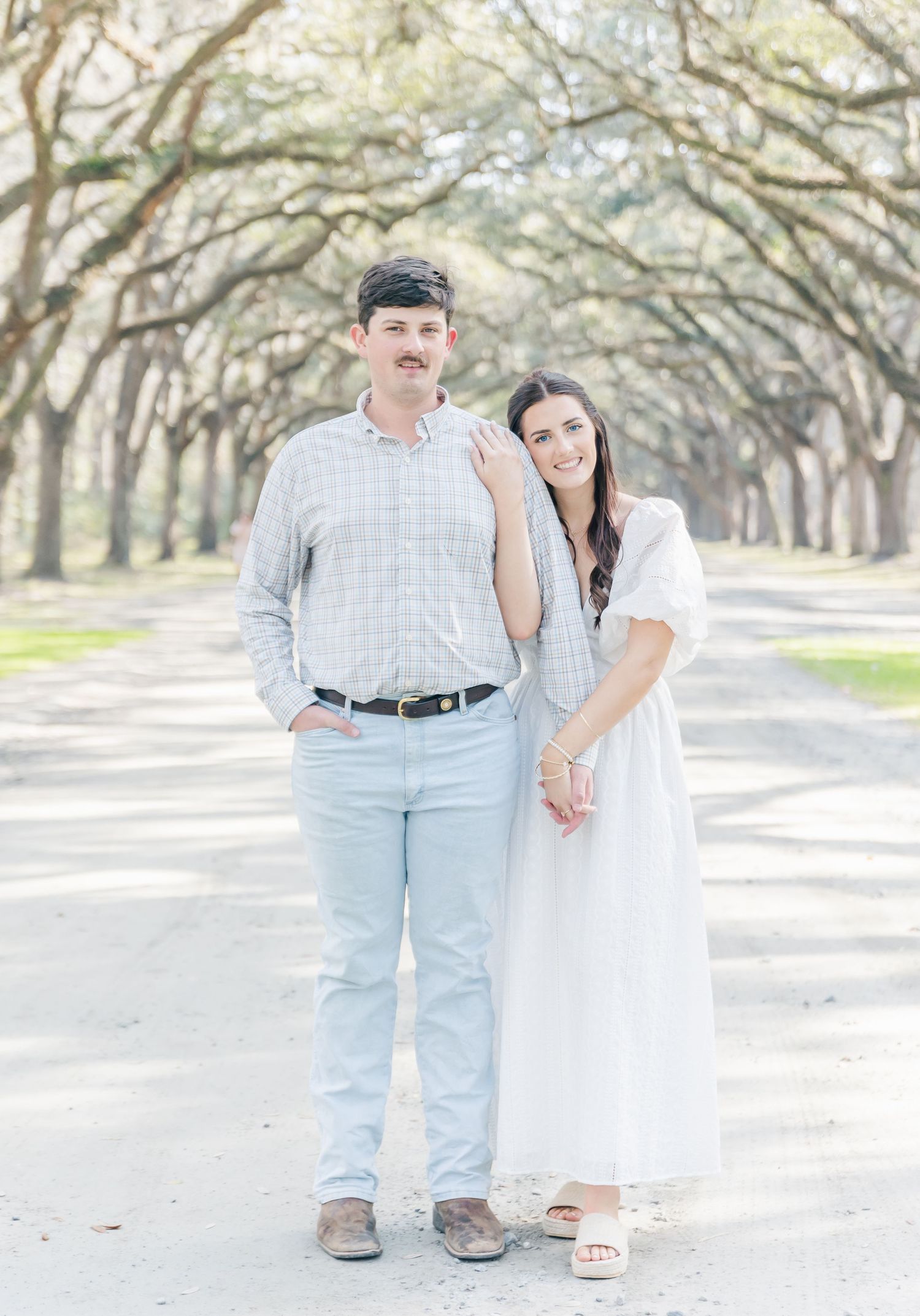 A couple holds hands while posing on a beautiful tree-lined avenue.