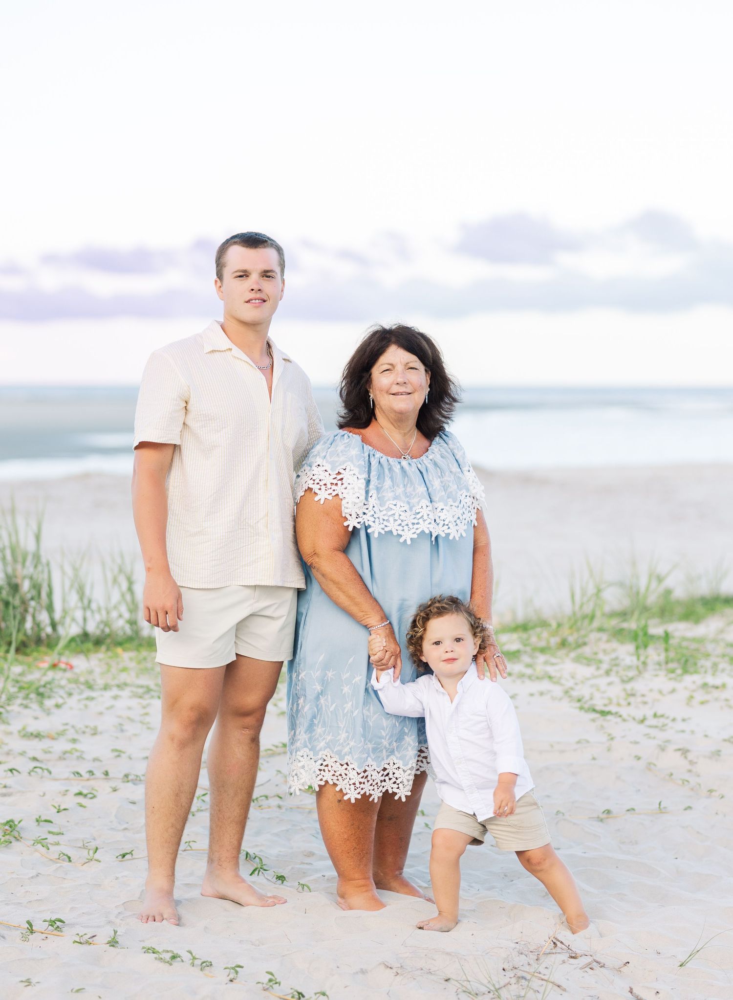 A family of three poses together on a sandy beach at dusk wearing light summer clothing.