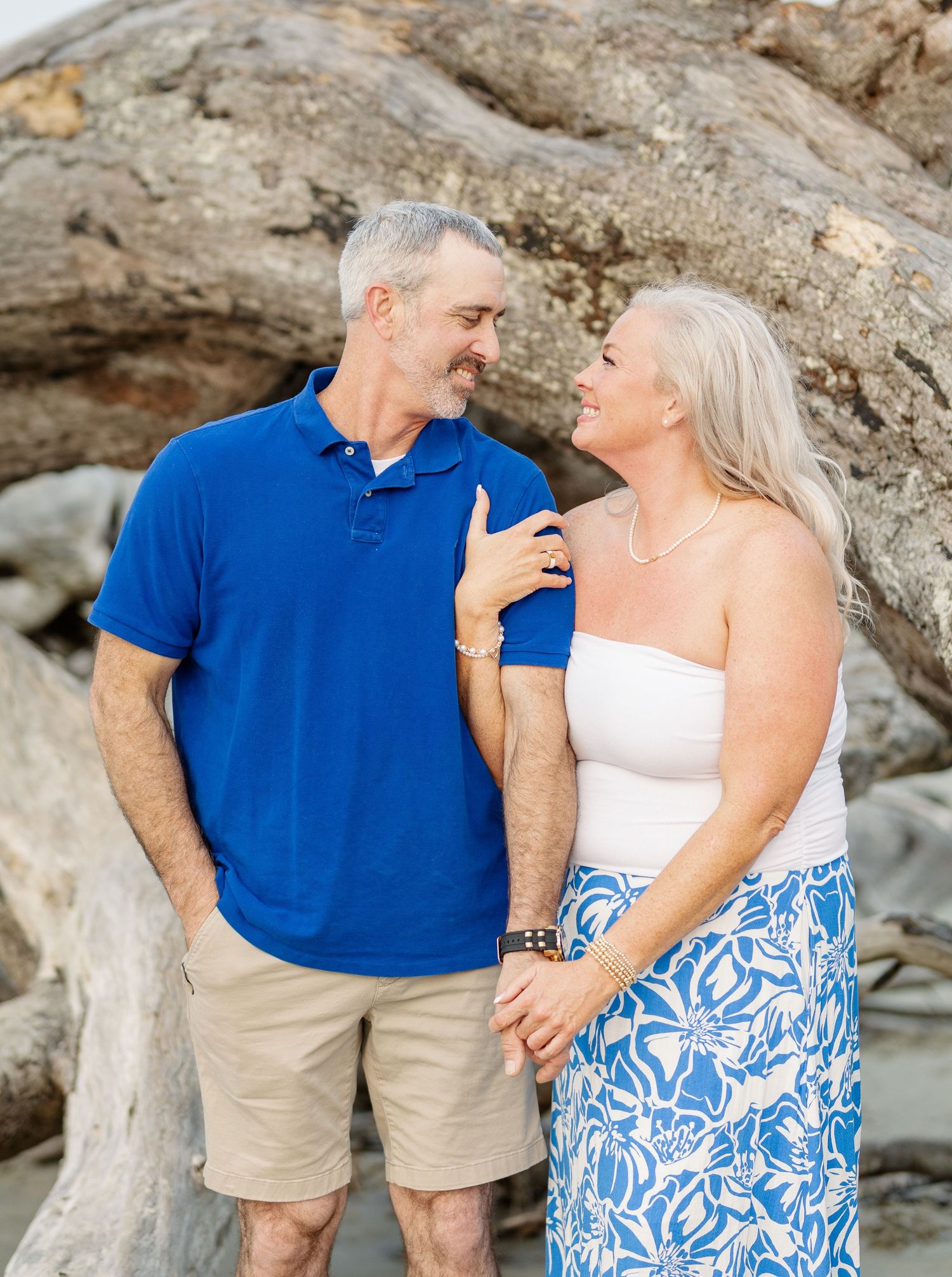 Happy mature couple in coordinating blue and white outfits embracing on rocky beach during outdoor photo session.