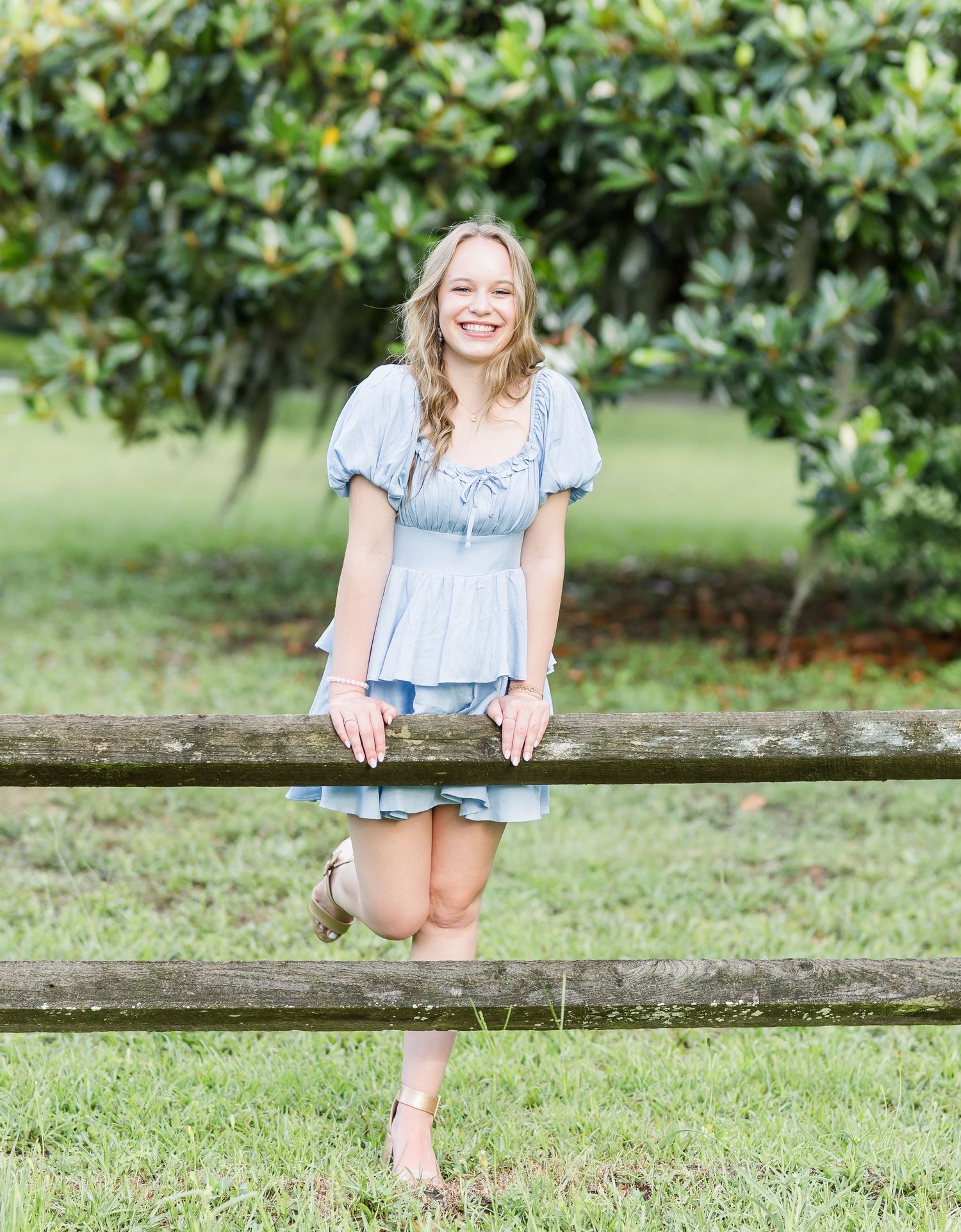 Someone wearing a light blue summer dress leans playfully on a wooden fence in a sunny garden setting.