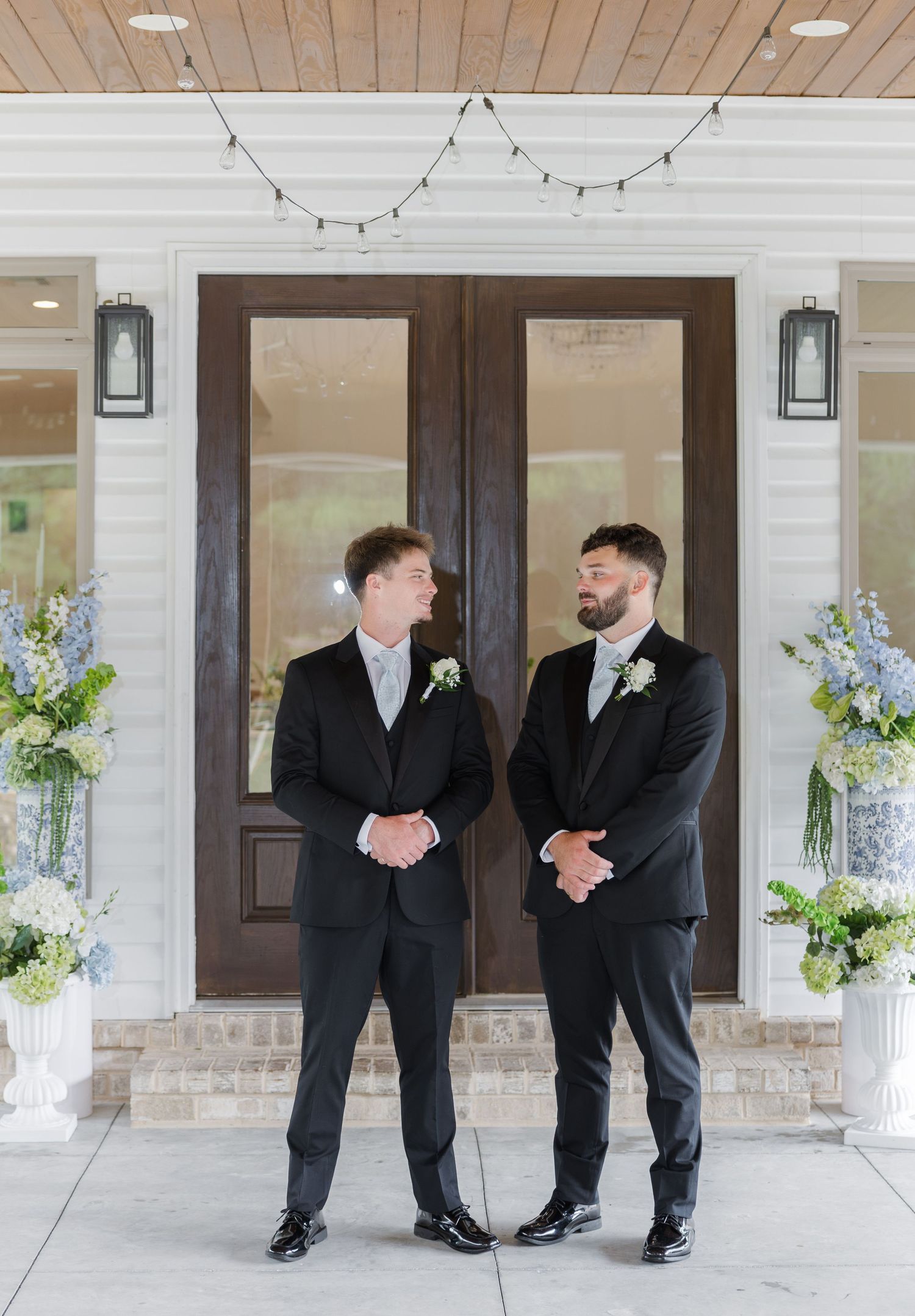Two groomsmen in black suits smiling at each other outside a venue with blue and white floral arrangements.