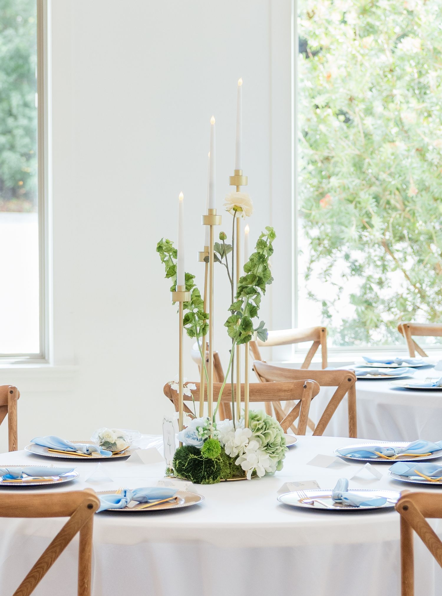 Elegant wedding tablescape with tall candles, greenery centerpiece, blue napkins, and wooden cross-back chairs in a bright room.