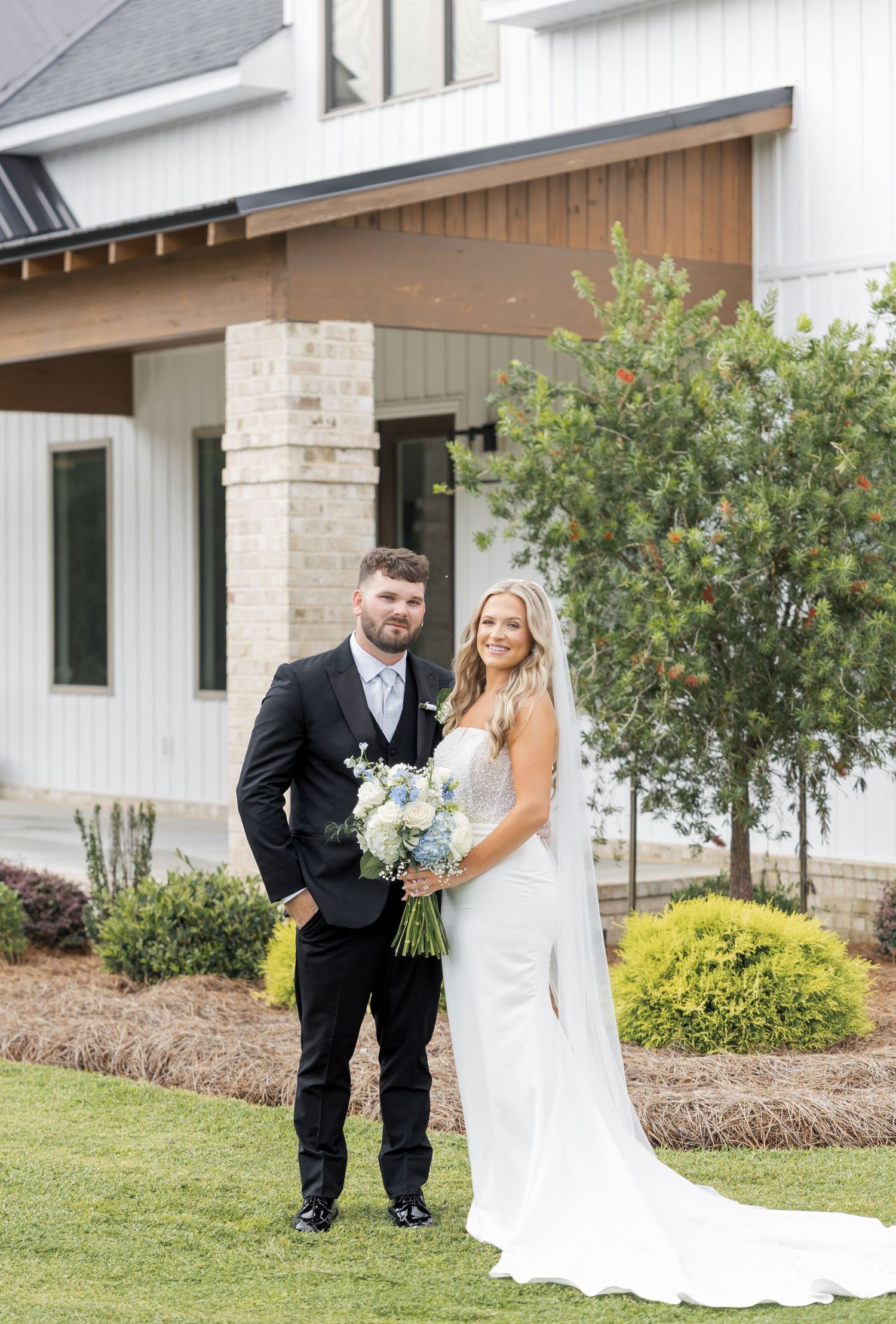 Bride and groom pose outside a white farmhouse venue, bride holding a blue and white floral bouquet.