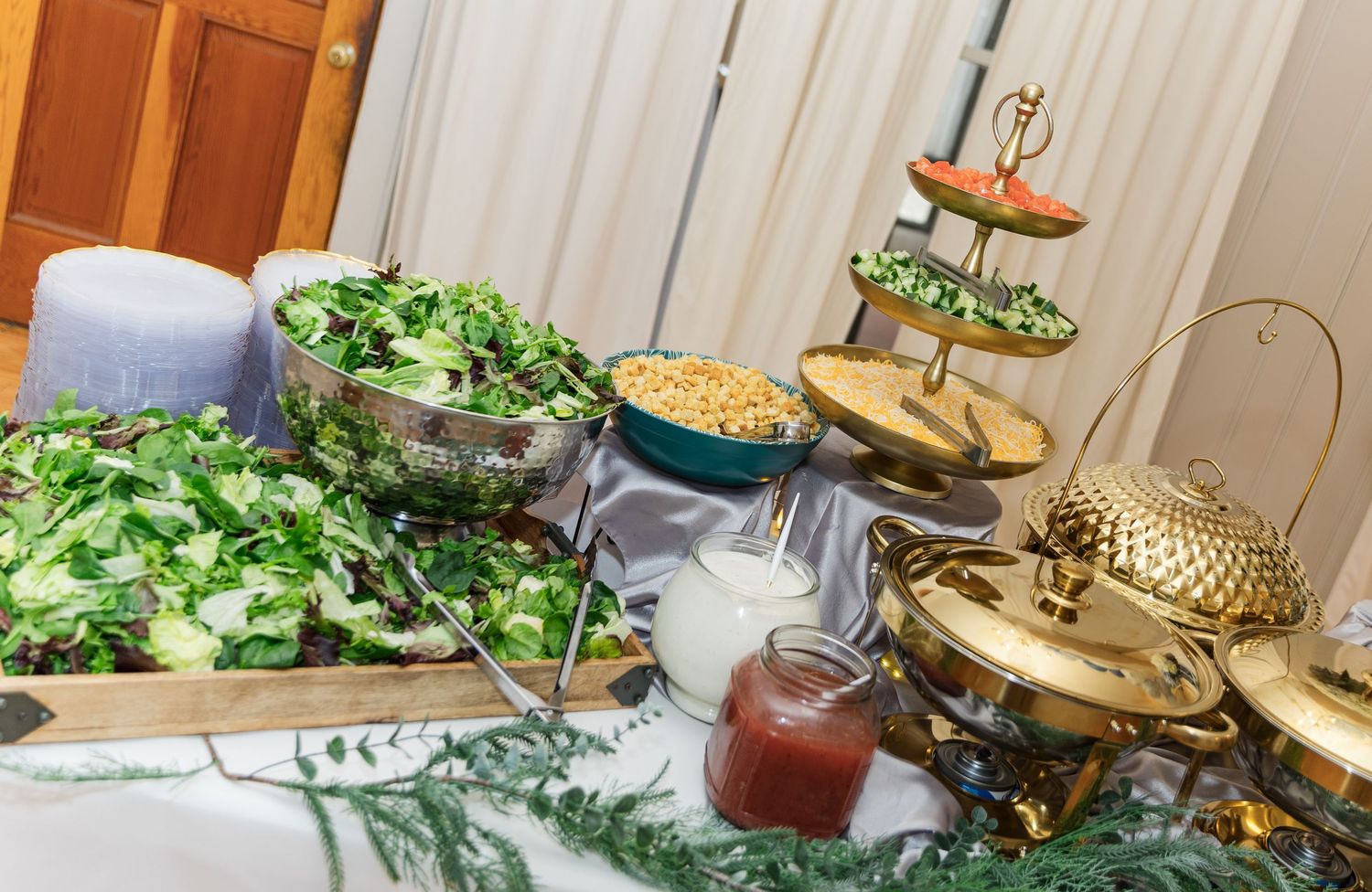 Elegant buffet table with fresh green salads, corn, chafing dishes, and a tiered appetizer stand at a catered event.