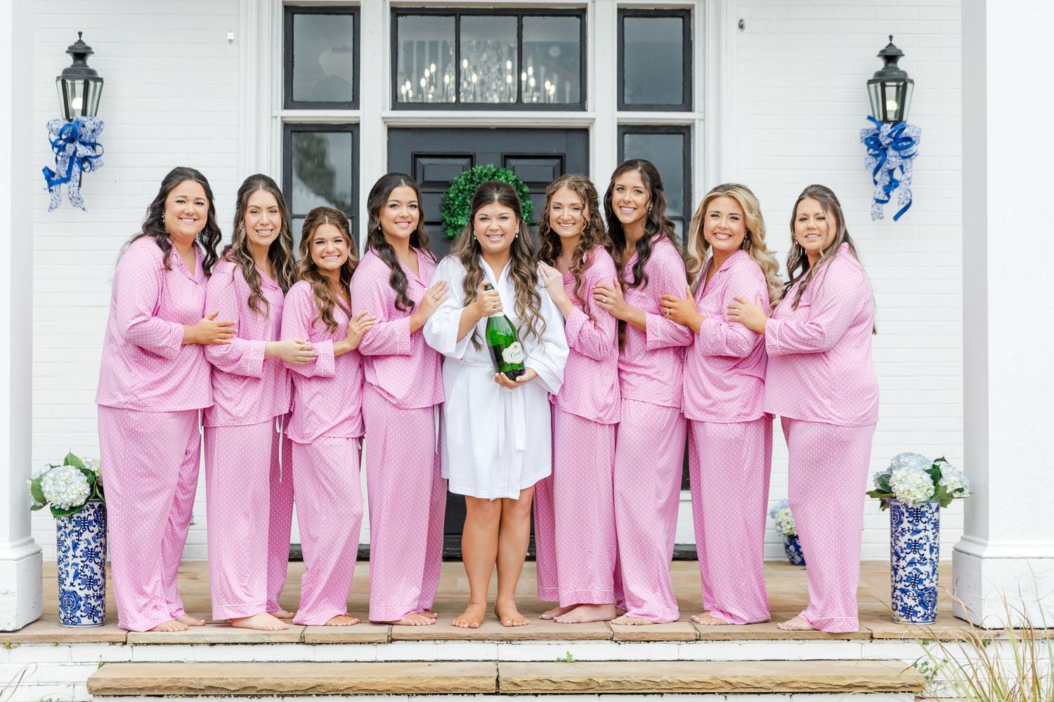 Bride in white robe holding champagne with bridesmaids in pink pajamas outside venue.